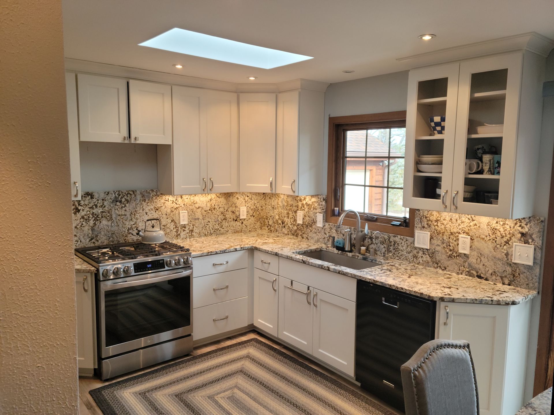 Bright kitchen with white cabinets, granite countertops, stainless stove, and a skylight above a striped rug.