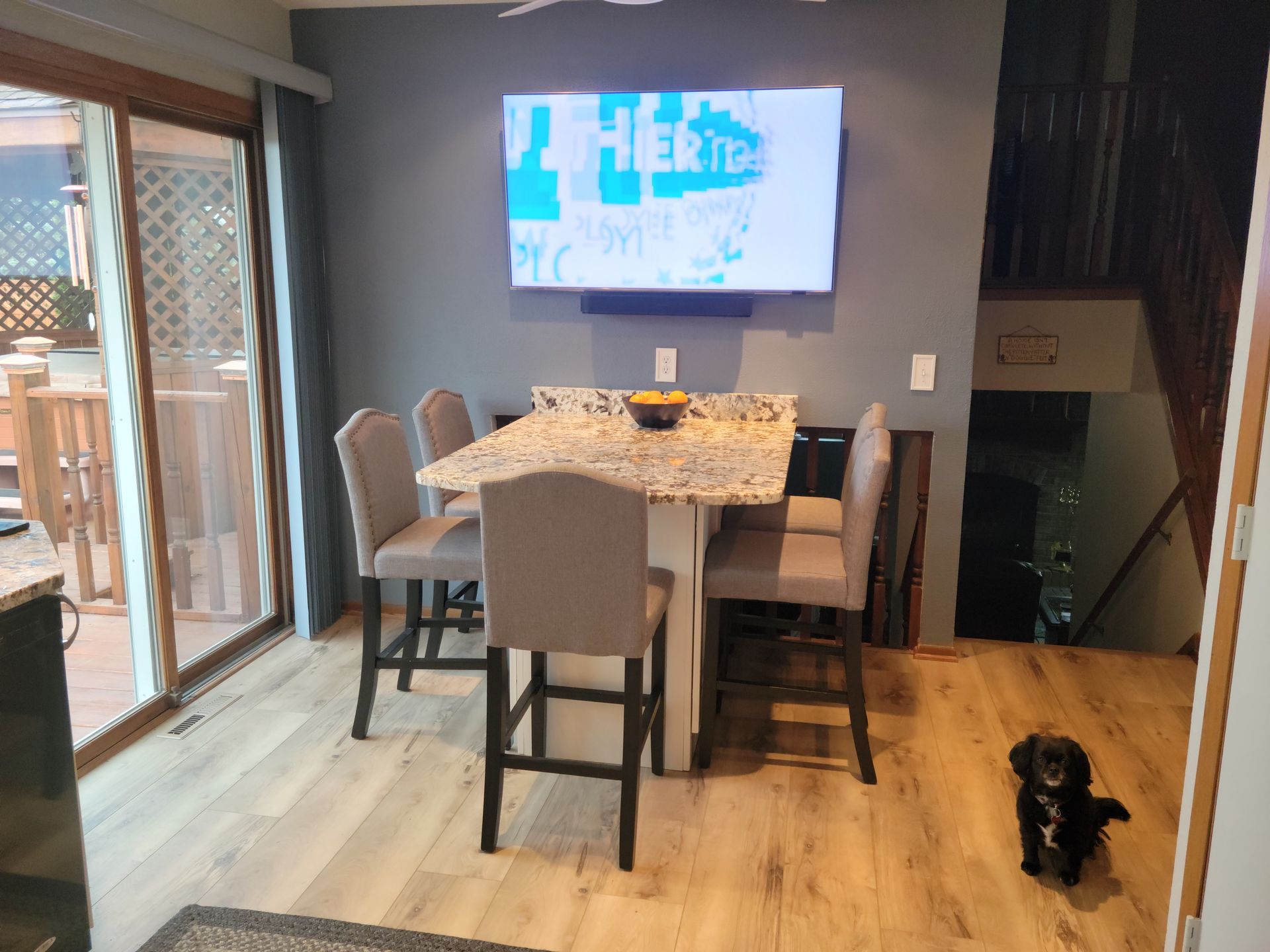 Modern dining room with bar stools, wall TV, open patio door, and a small dog on the floor