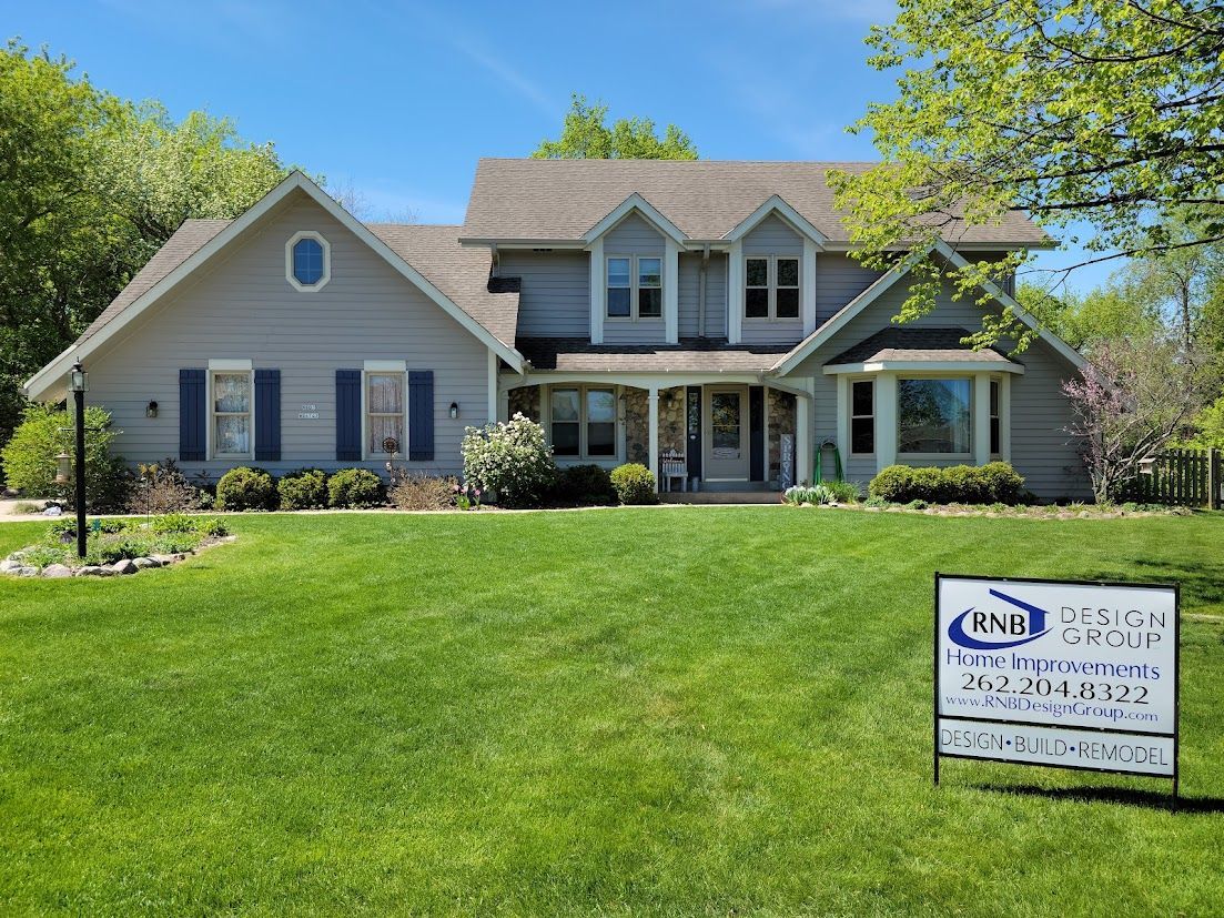 Gray house with blue shutters and porch, viewed across a green lawn under a clear sky.