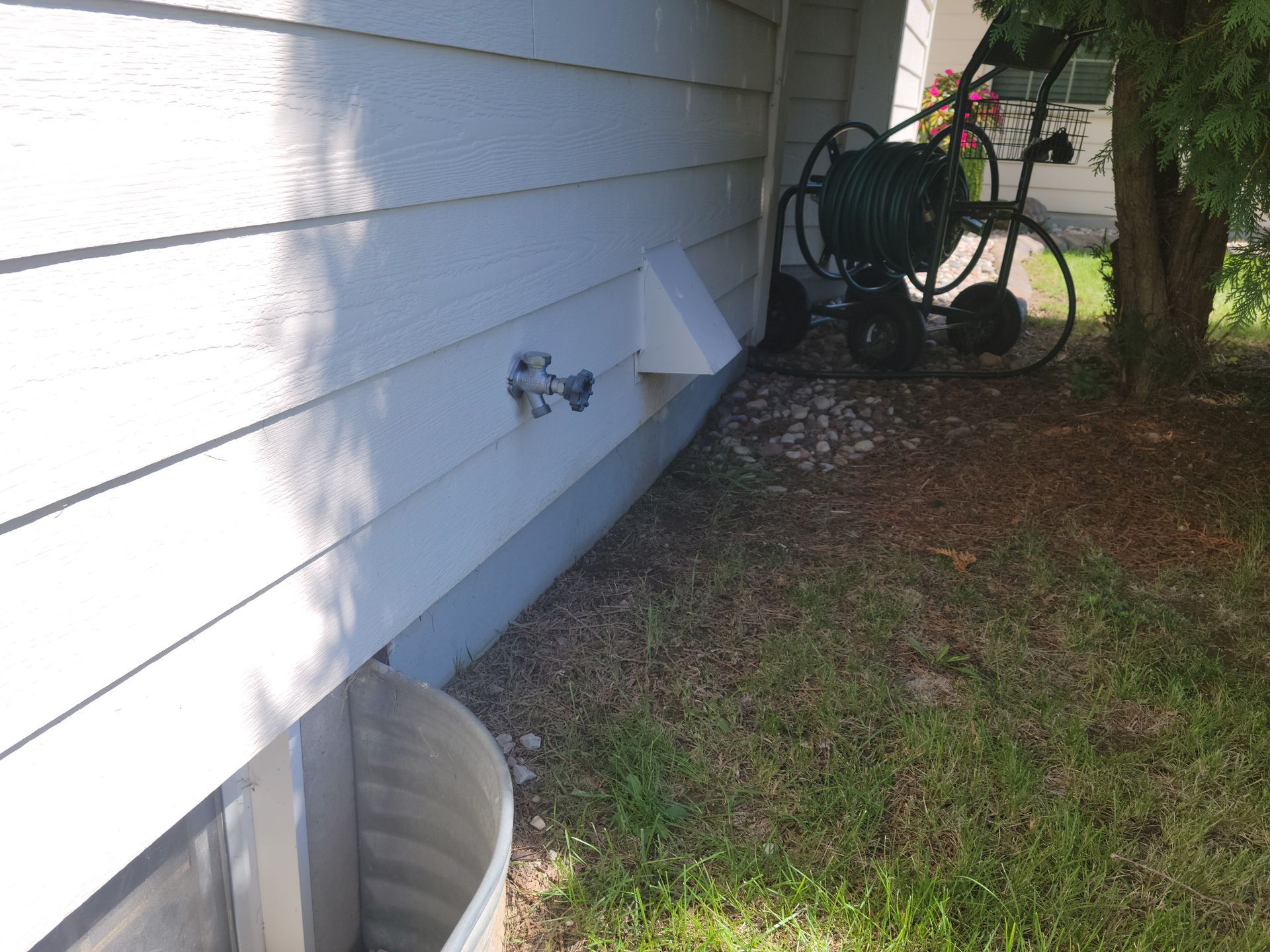 Side of a white house with a wall spigot, window well, and garden hose reel near grass and trees