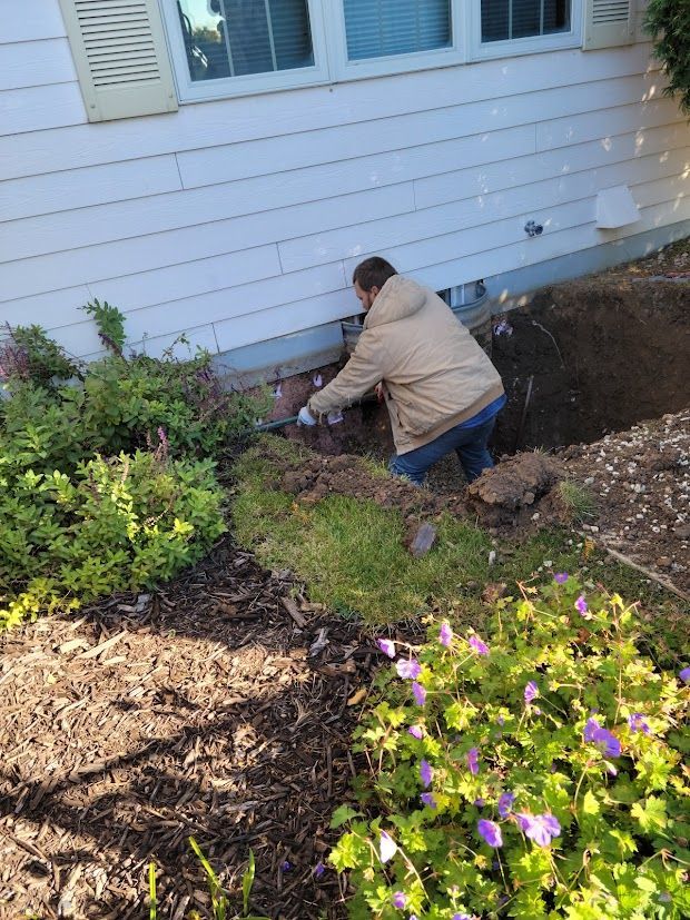 Person kneeling by a house foundation, digging in a garden bed beside shrubs and flowers.
