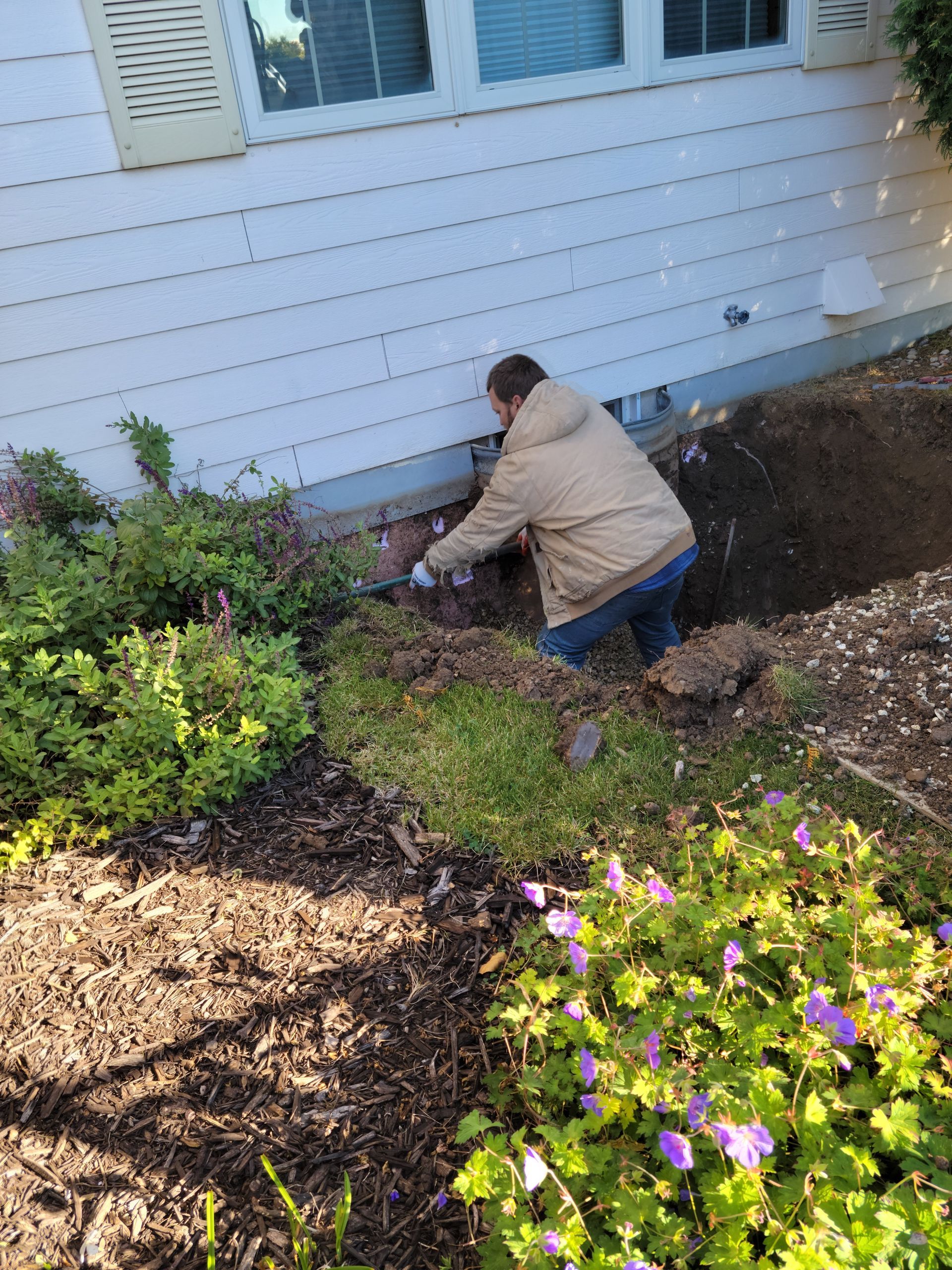 Person kneeling in a garden bed near a house, planting or weeding among flowers and mulch.