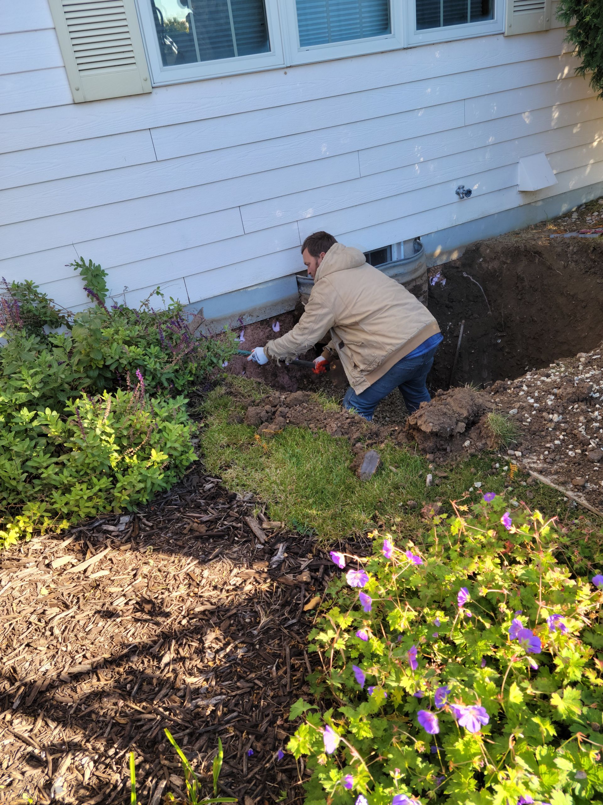 Person kneeling in a garden bed beside a white house, planting or digging near shrubs and flowers