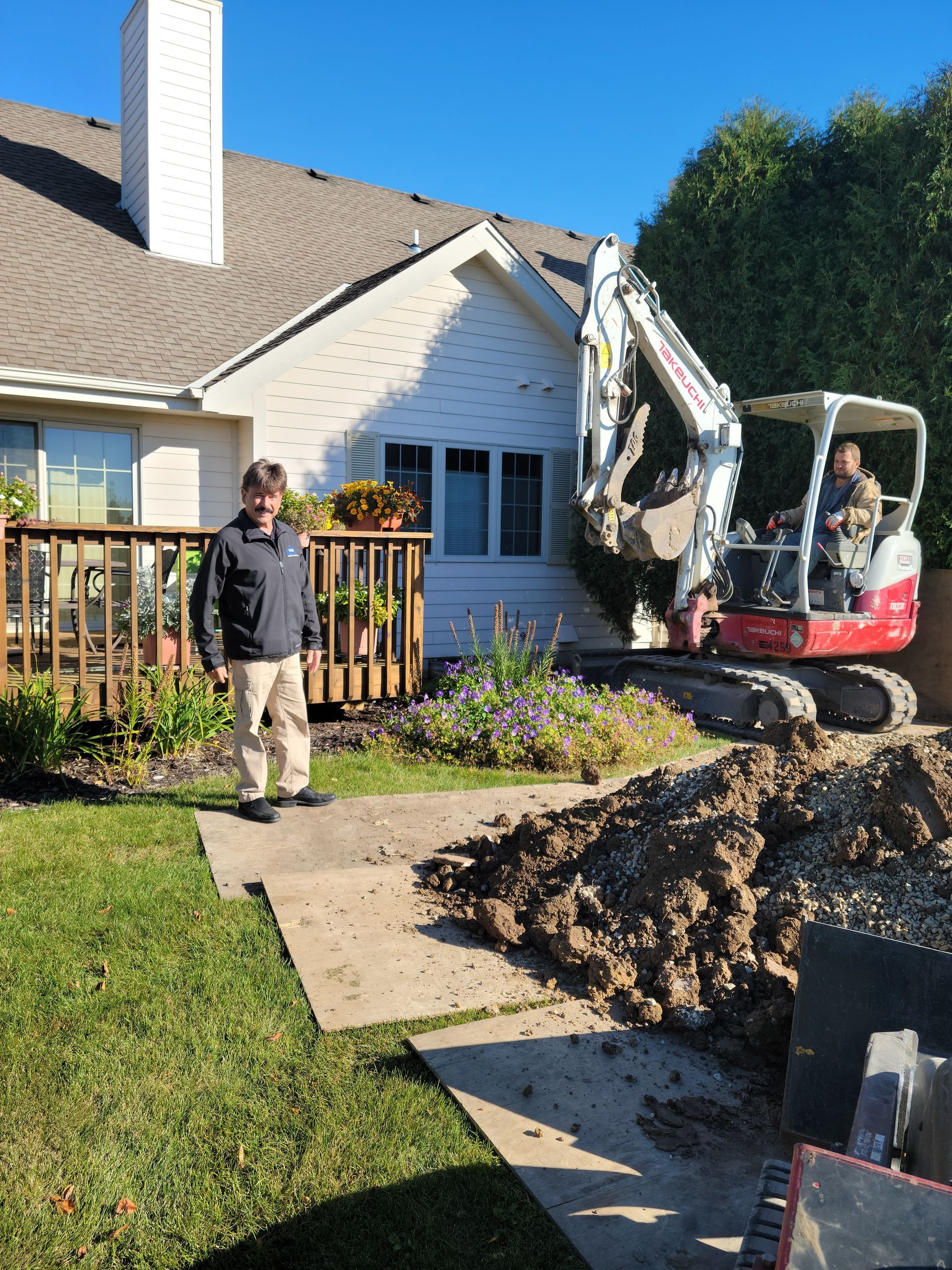 Mini excavator digging in front of a white house while a person stands nearby on a sunny day