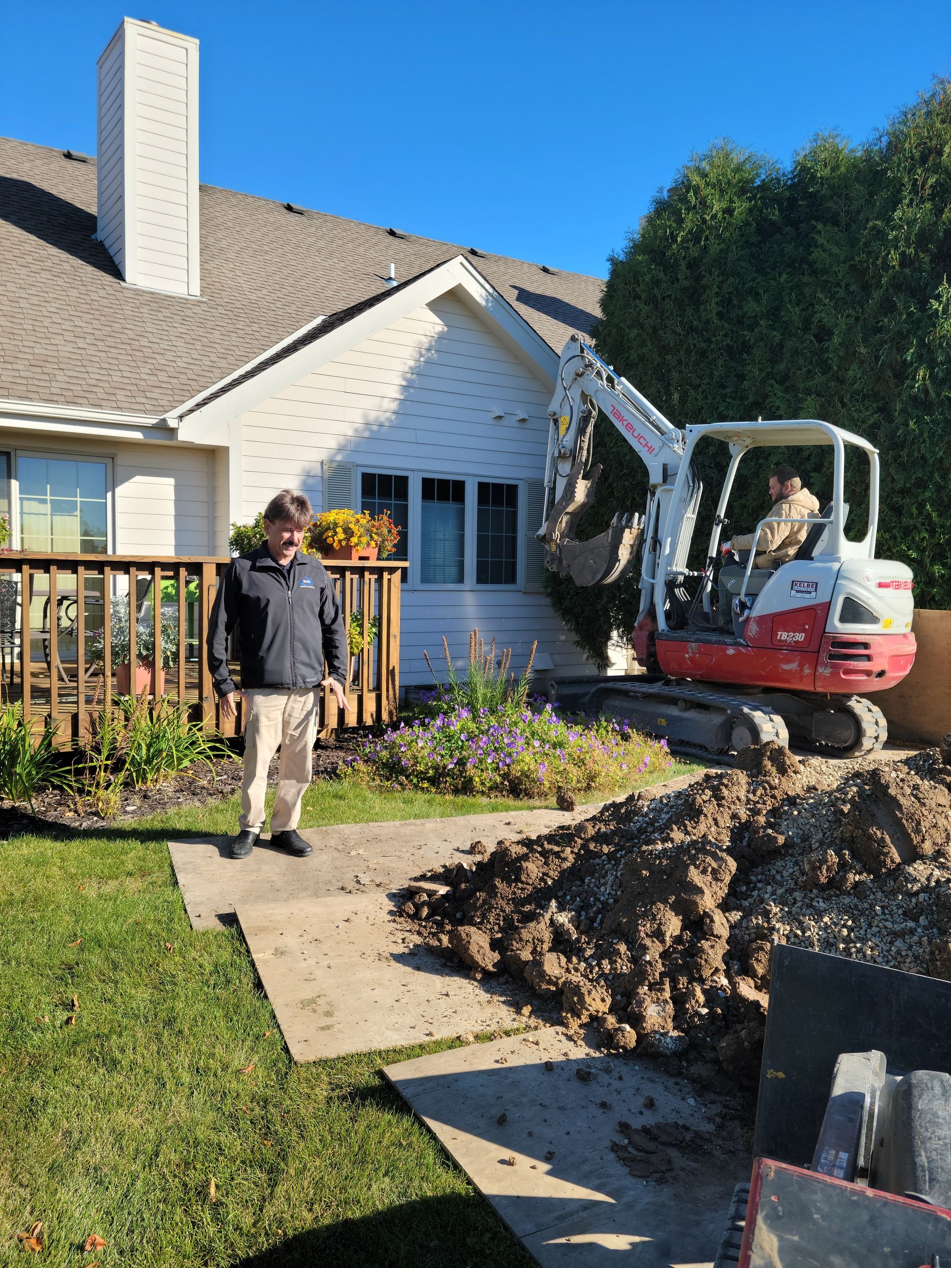Person stands by a house as a mini excavator digs up soil near a walkway and deck.