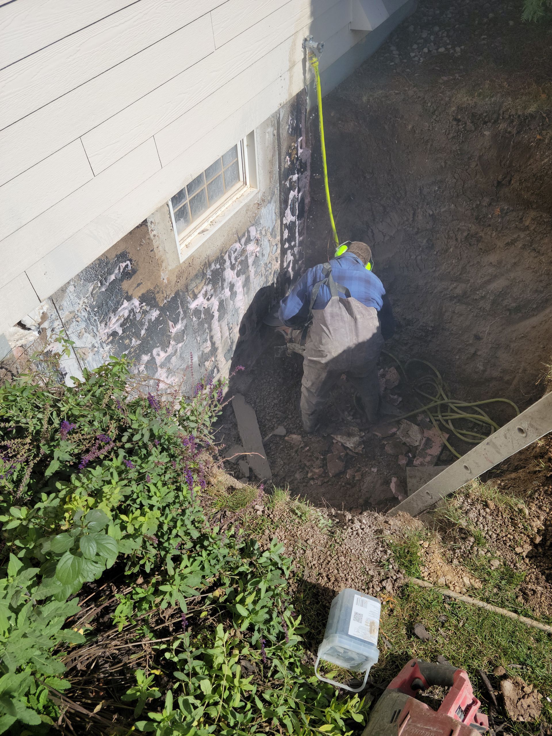 Worker in blue jacket and yellow harness in a dug hole beside a house foundation.