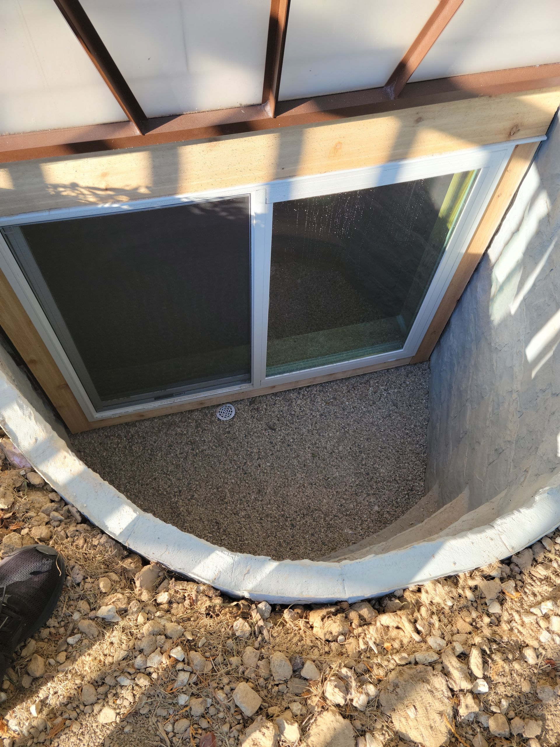 Basement window well with white trim, gravel floor, and two dark window panes under a wooden structure