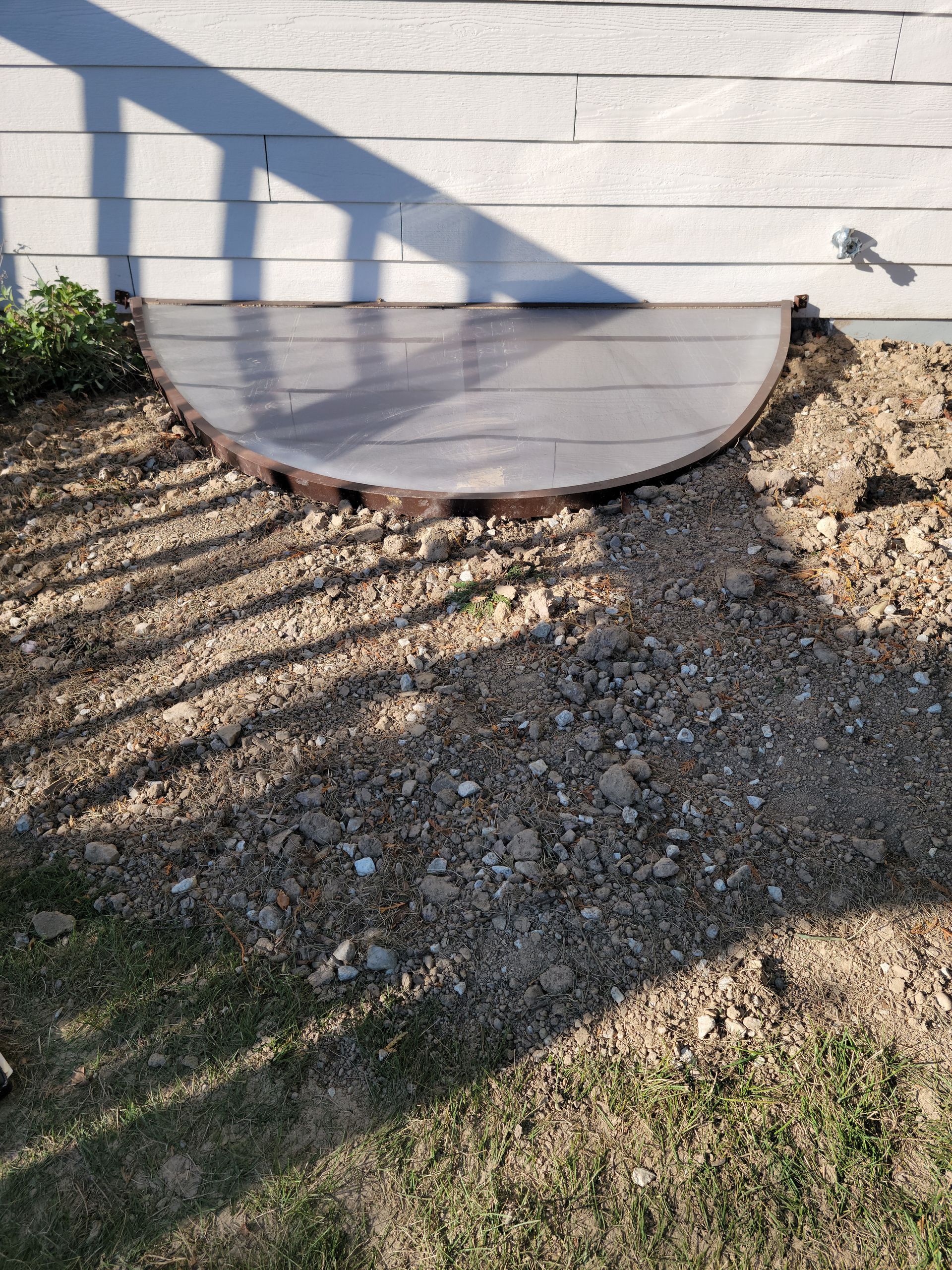 Curved window well cover beside a house, with gravel and striped shadows from a railing.