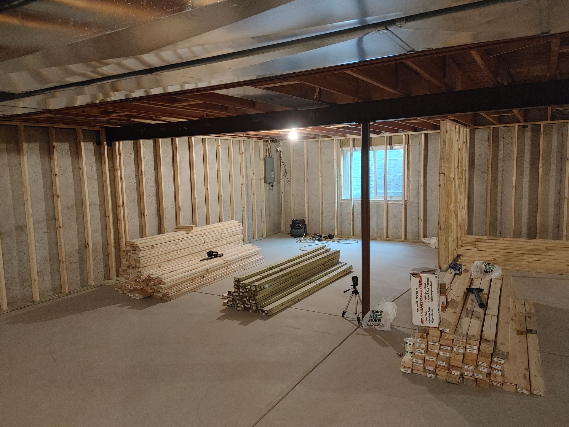 Unfinished basement framing with insulation, exposed beams, and stacks of lumber on the concrete floor.