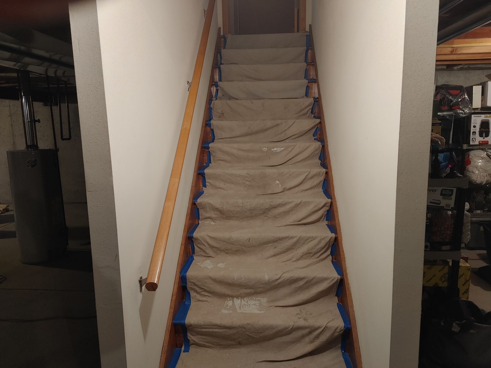 Narrow staircase covered with brown tarp, flanked by white walls and a wooden handrail in a basement.