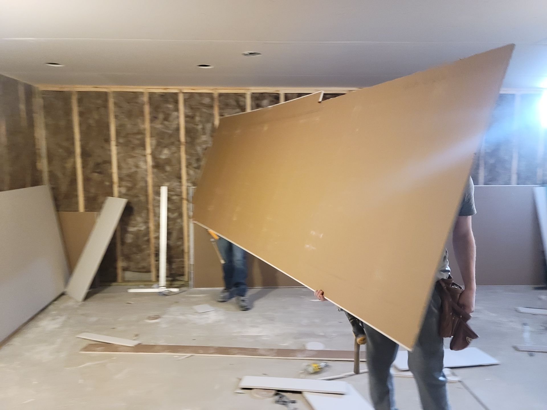 People carrying a large beige drywall panel in an unfinished room under construction
