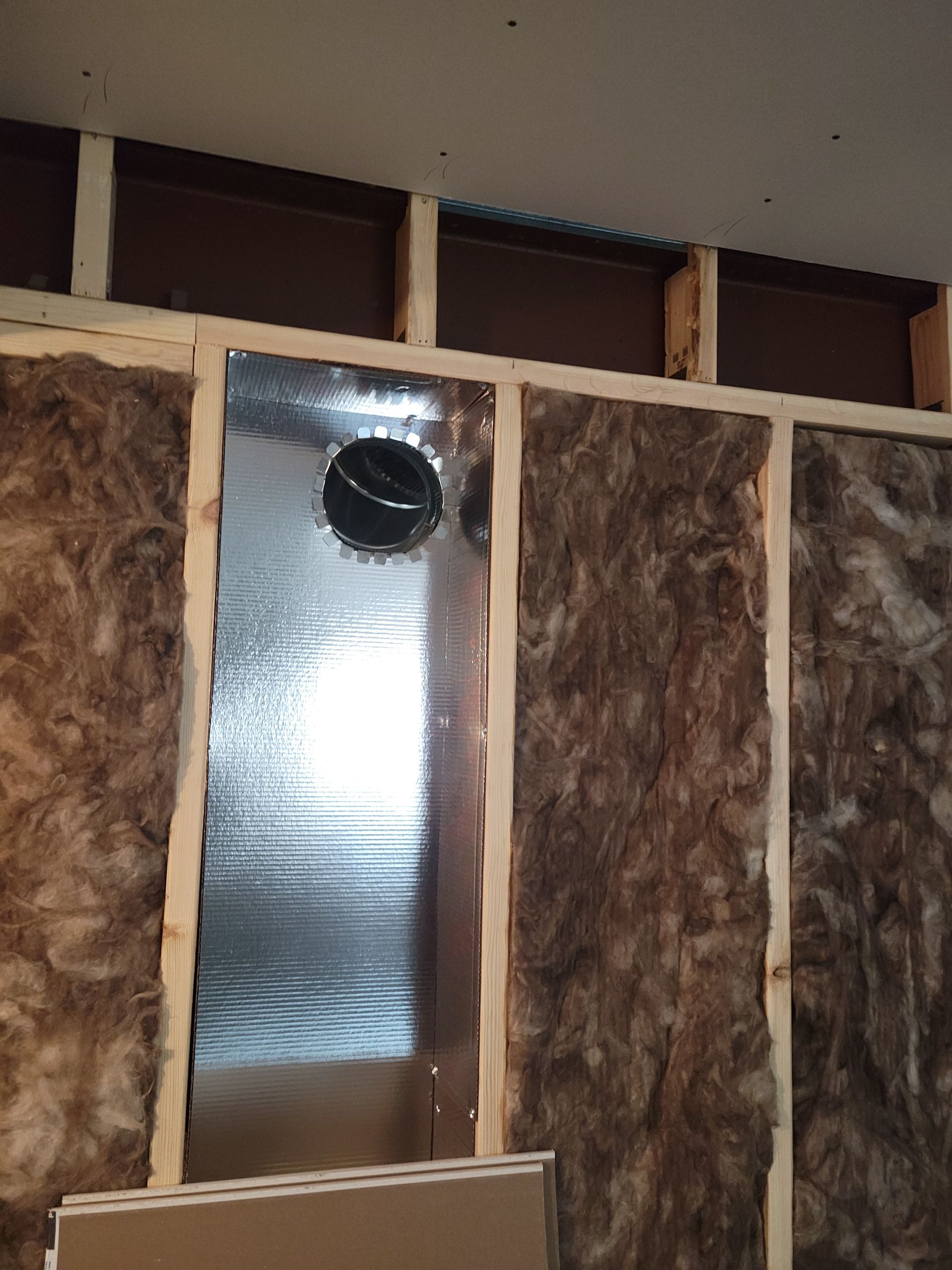Unfinished wall with exposed insulation, foil ducting, and a round black vent opening.