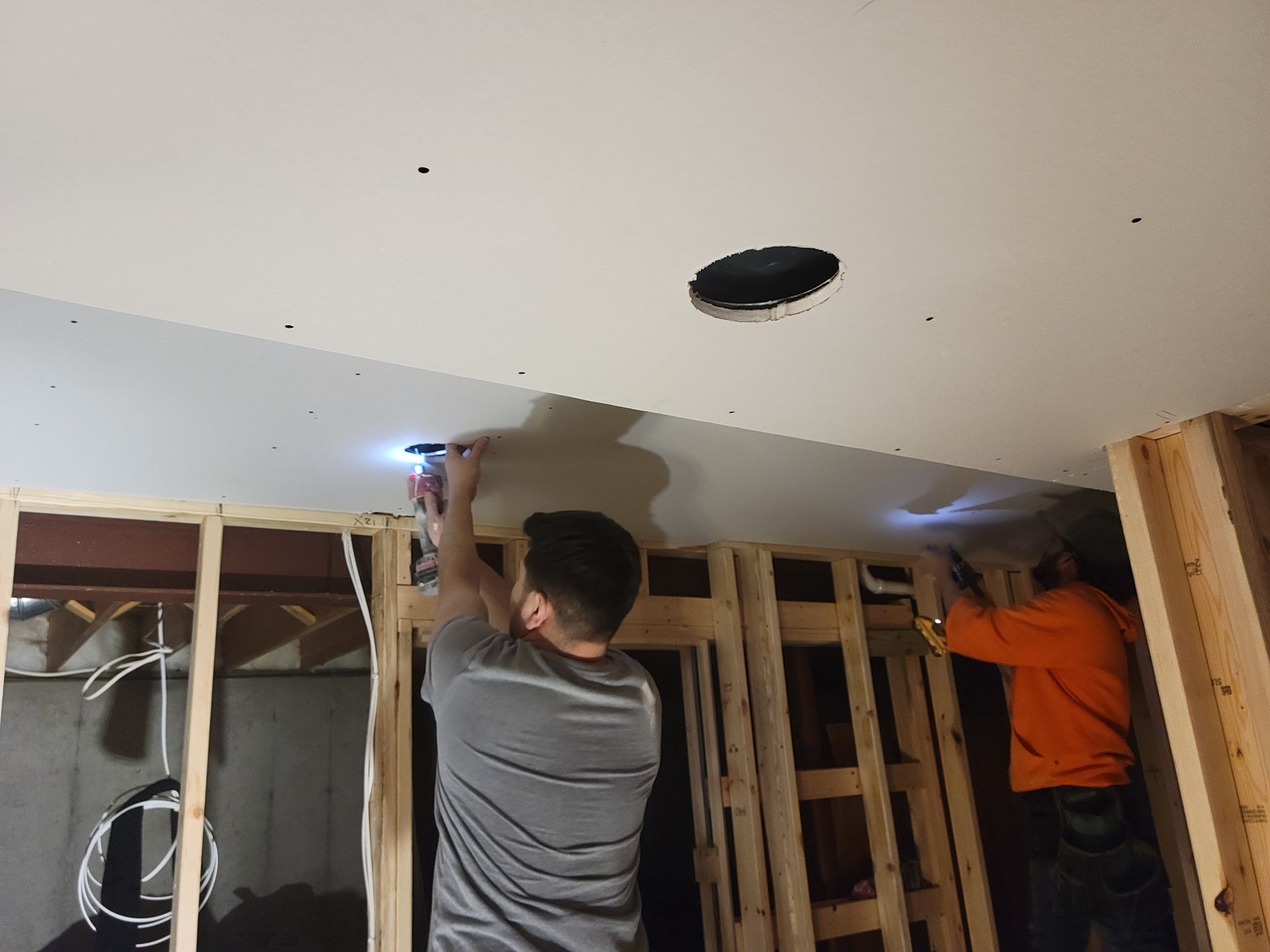 Two workers installing drywall in an unfinished room with exposed framing and ceiling cutouts.