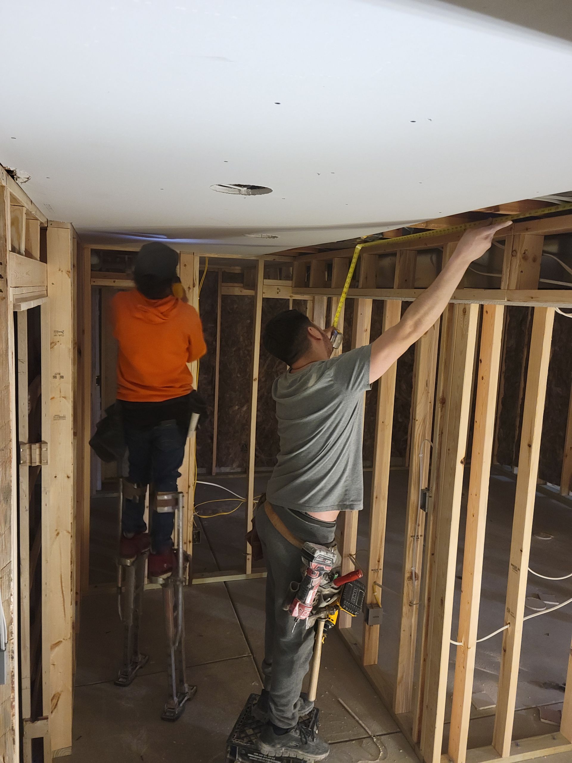 Two workers installing drywall framing inside a room under construction, one on a ladder.
