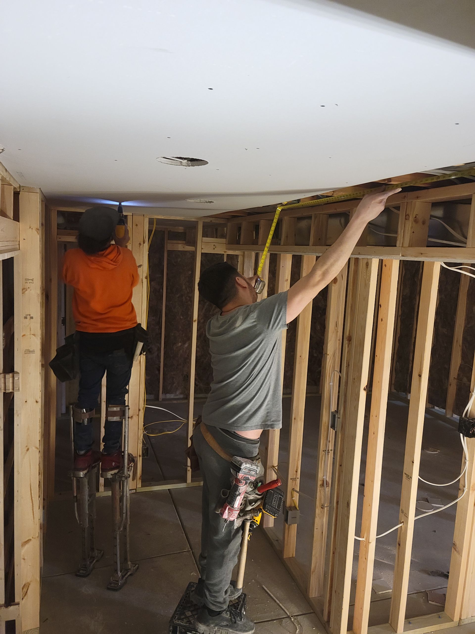 Two workers framing a wall during home renovation, one standing on a ladder and one reaching overhead.