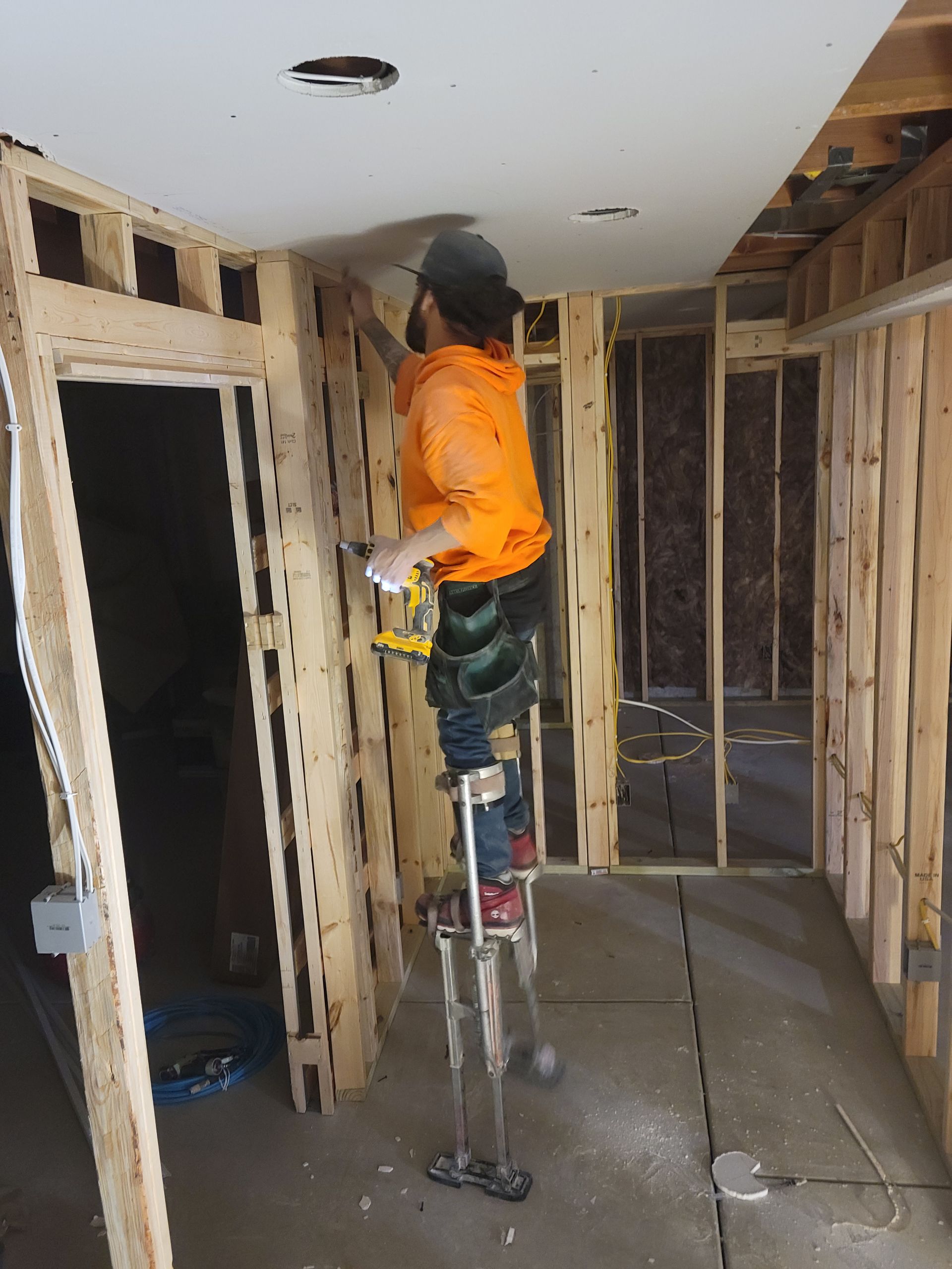 Worker on a ladder installing drywall in an unfinished room with exposed framing.