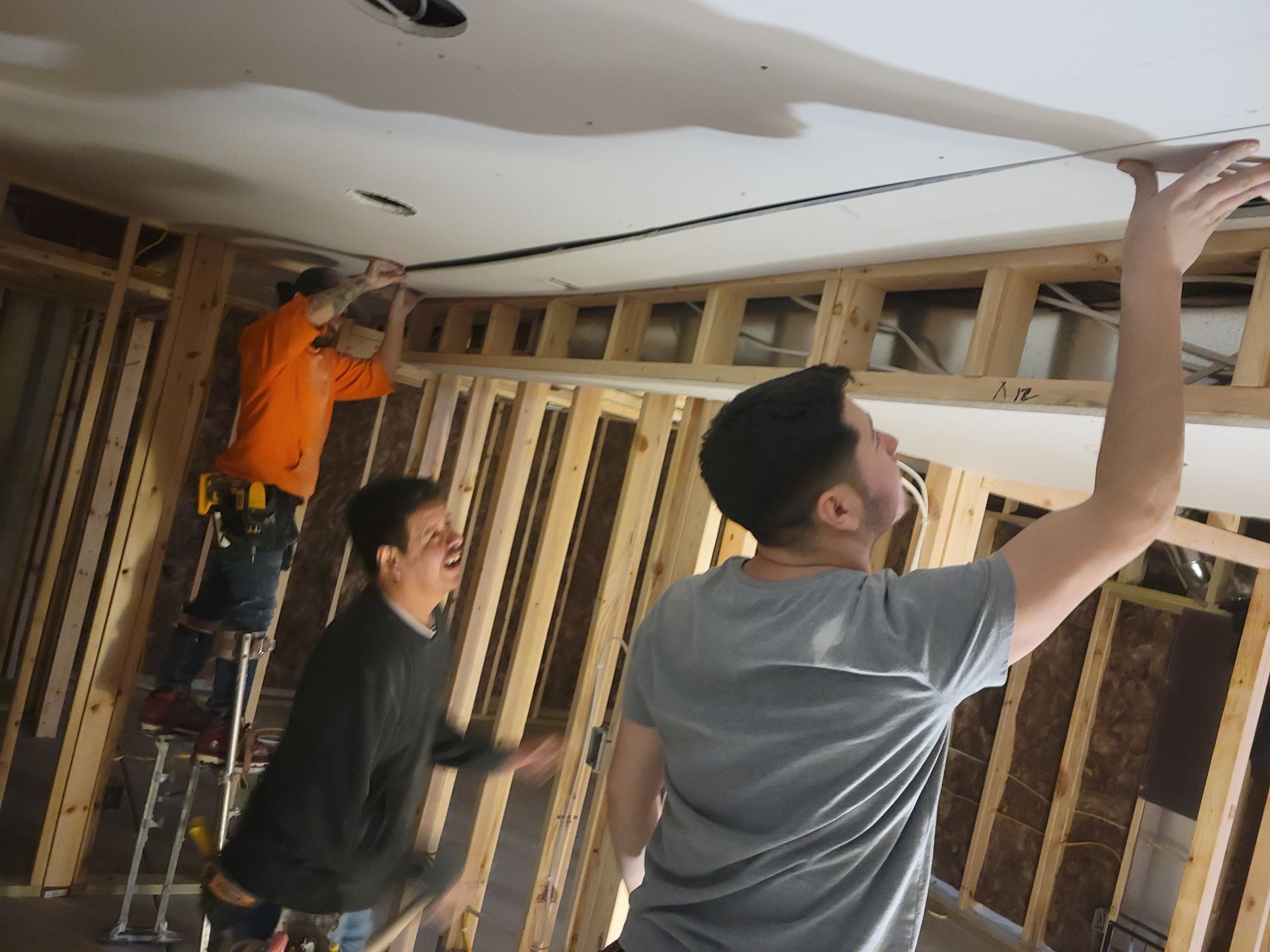 People installing drywall in a room with exposed studs and ceiling framing.