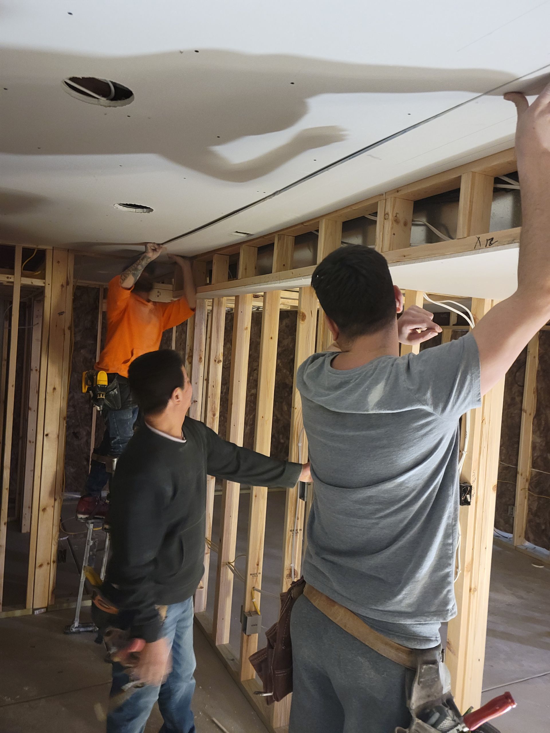 Two workers installing drywall and framing in an unfinished room with exposed studs and ceiling damage