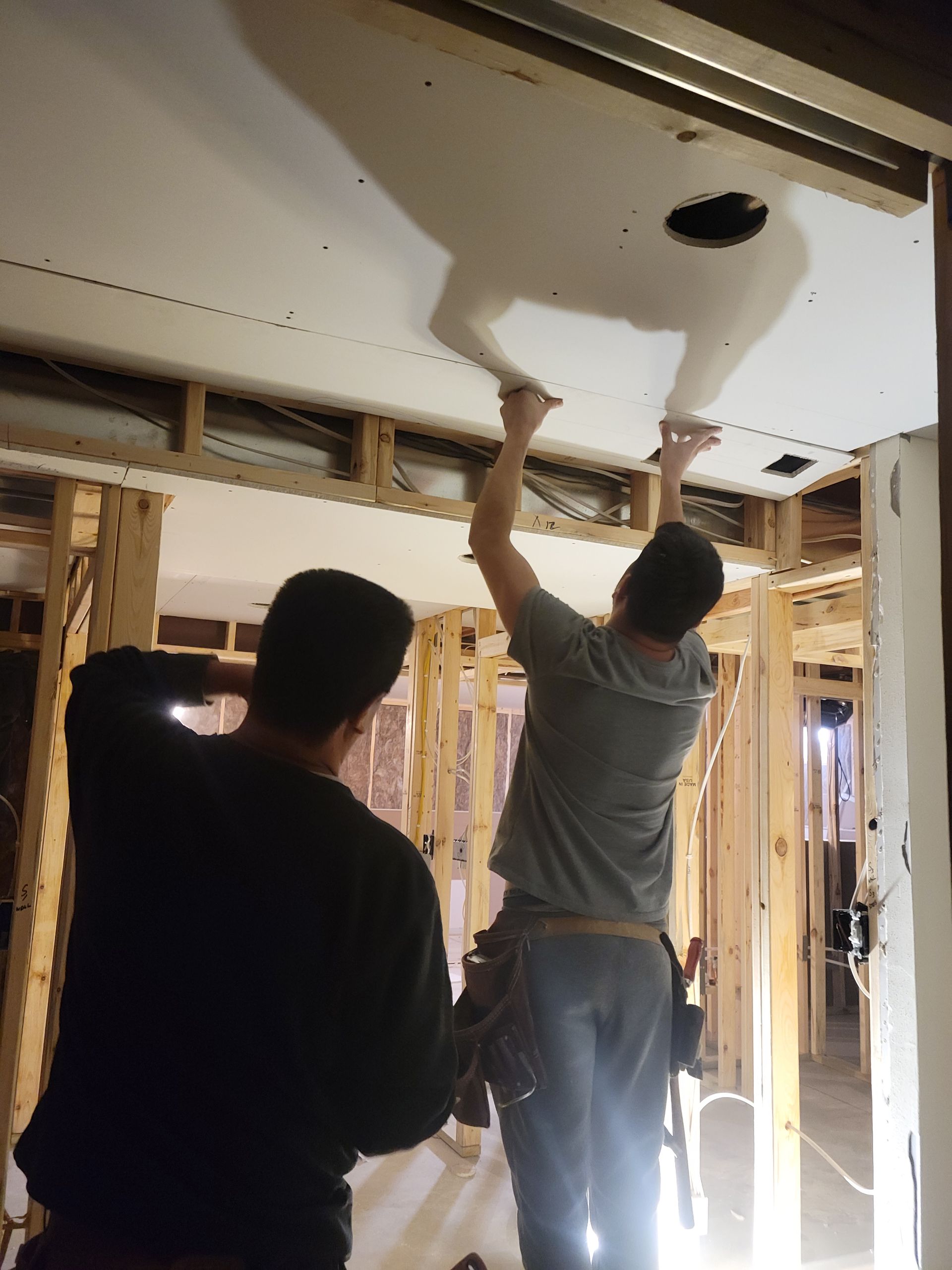 Two workers installing drywall on a ceiling in an unfinished room with exposed framing.