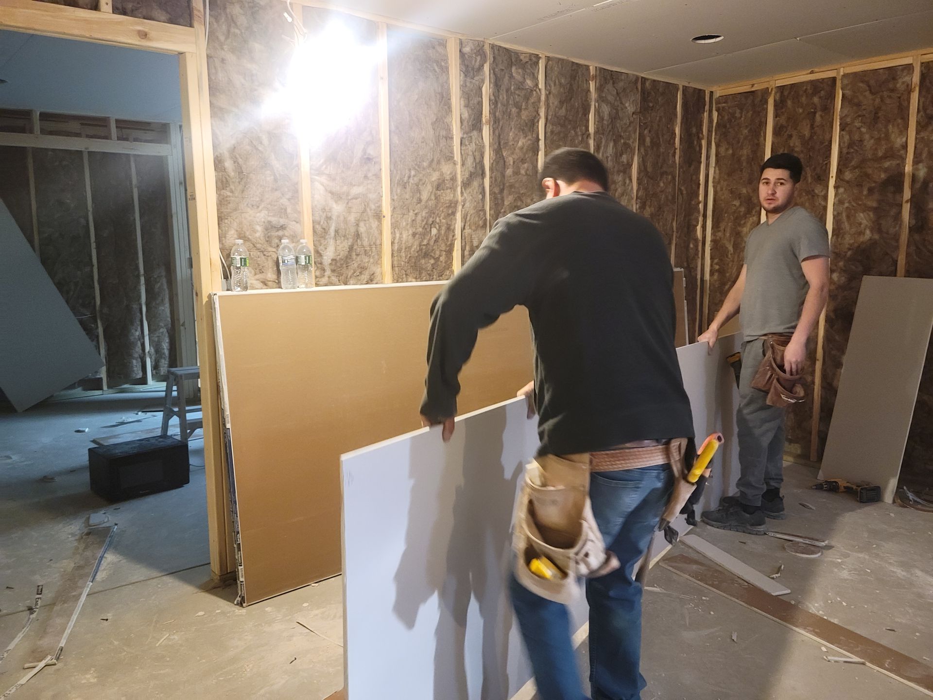 Two workers installing drywall in a partially finished room with exposed insulation and a bright work light