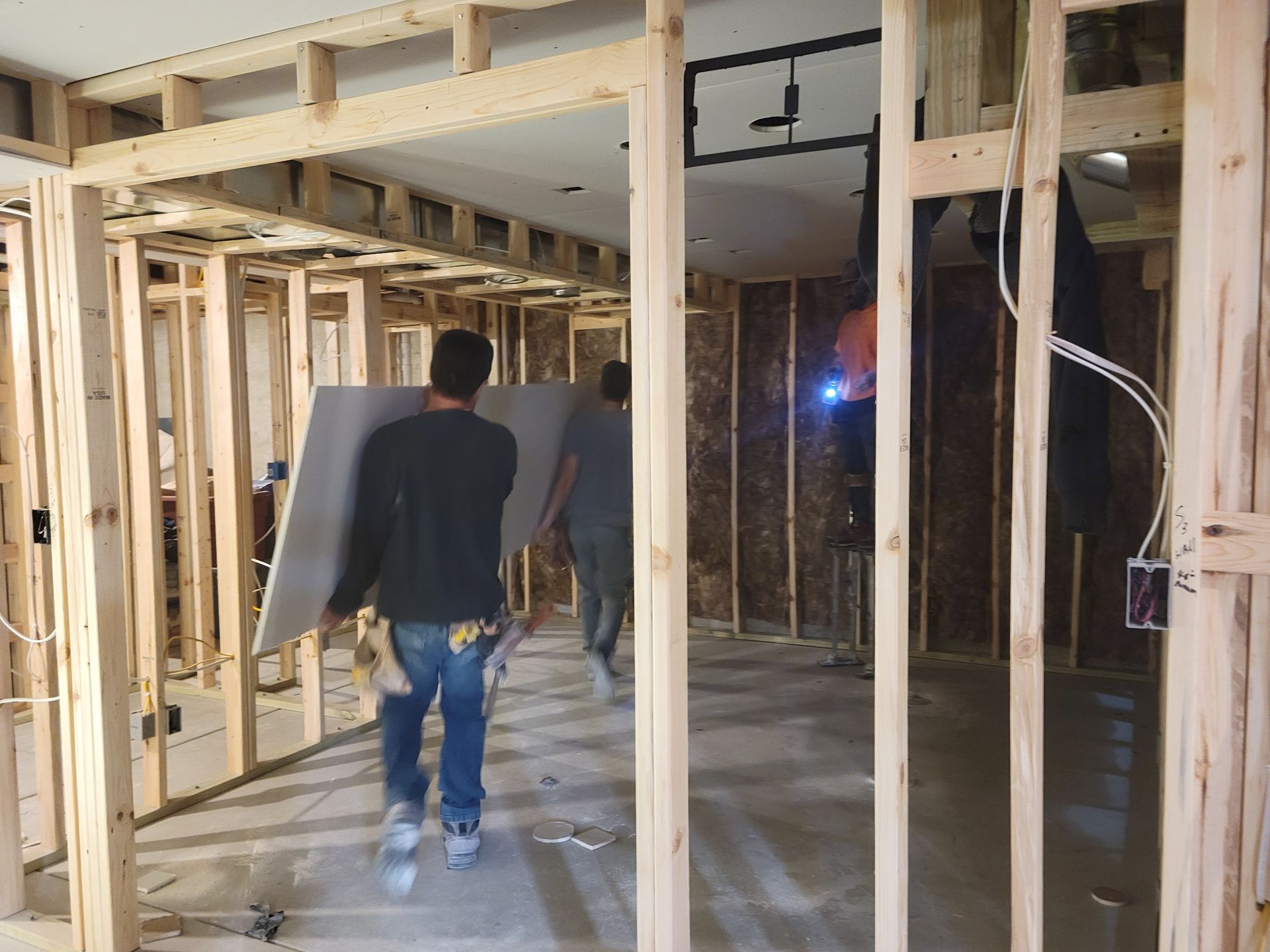 Construction site interior with exposed wood framing and two workers carrying drywall sheets