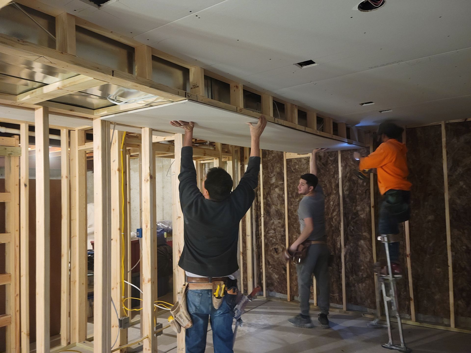 Workers framing a room interior, installing wood studs and ceiling beams during construction.