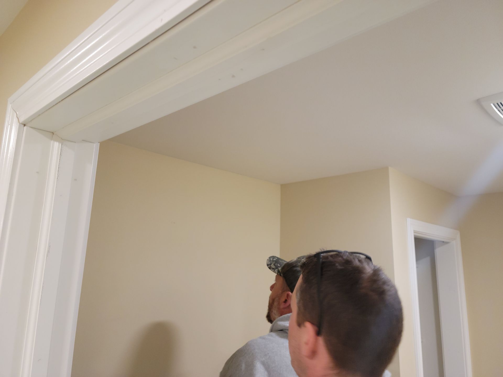 Two men inspecting a beige room corner beneath white trim near a doorway
