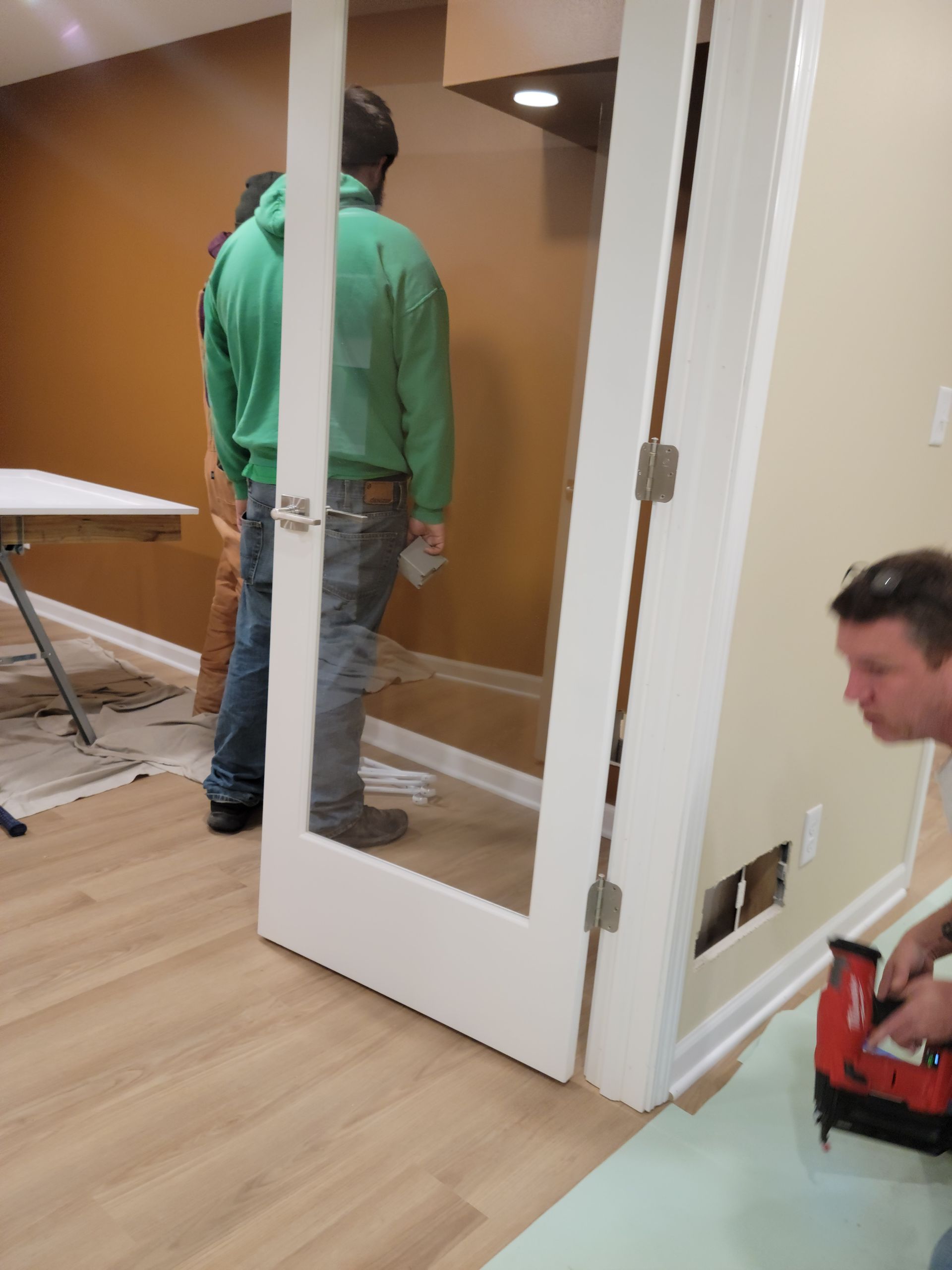 Two workers installing a glass door in a room with wood floors and beige walls.