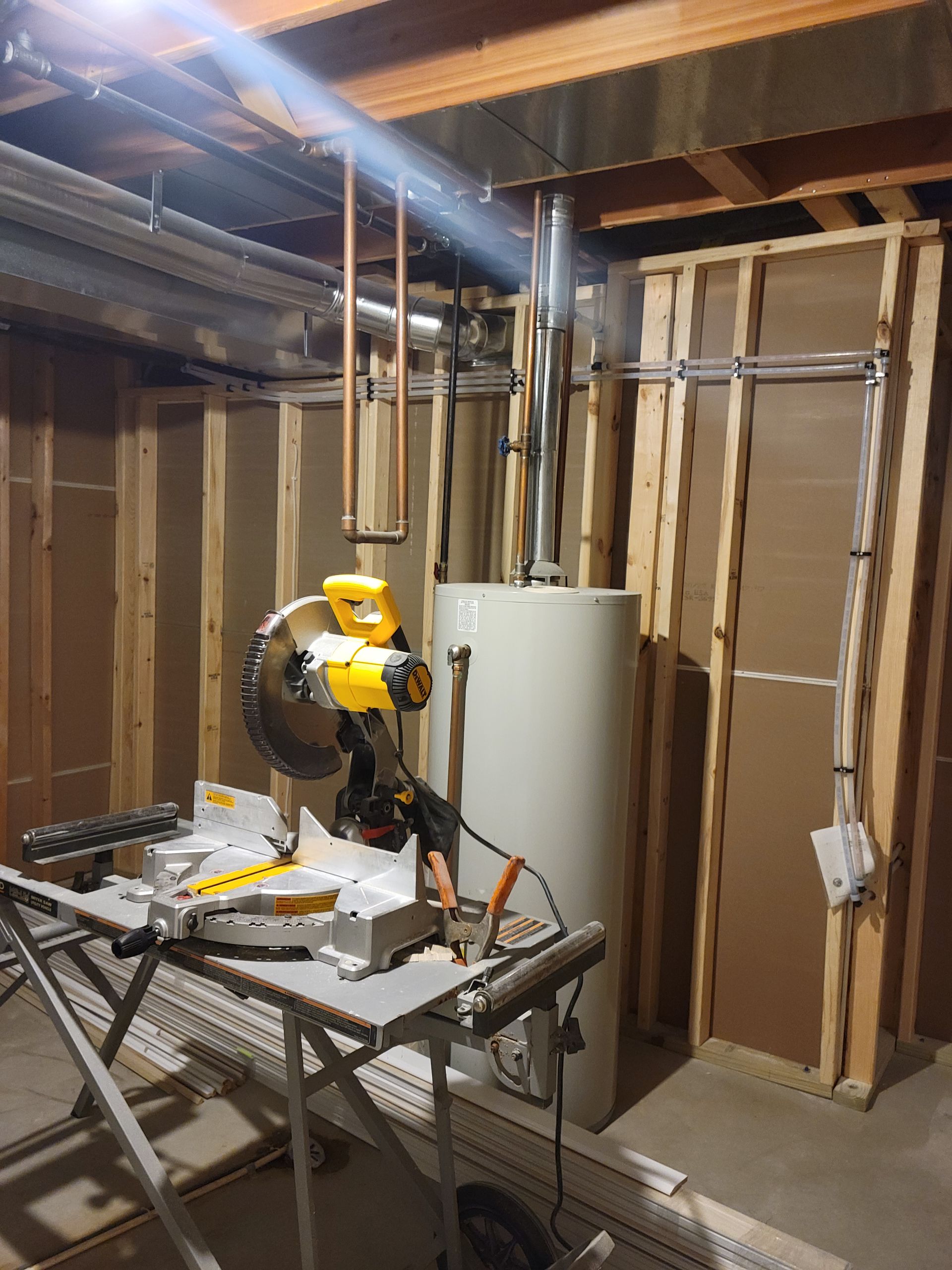 Yellow miter saw on workbench in unfinished basement with exposed studs and pipes