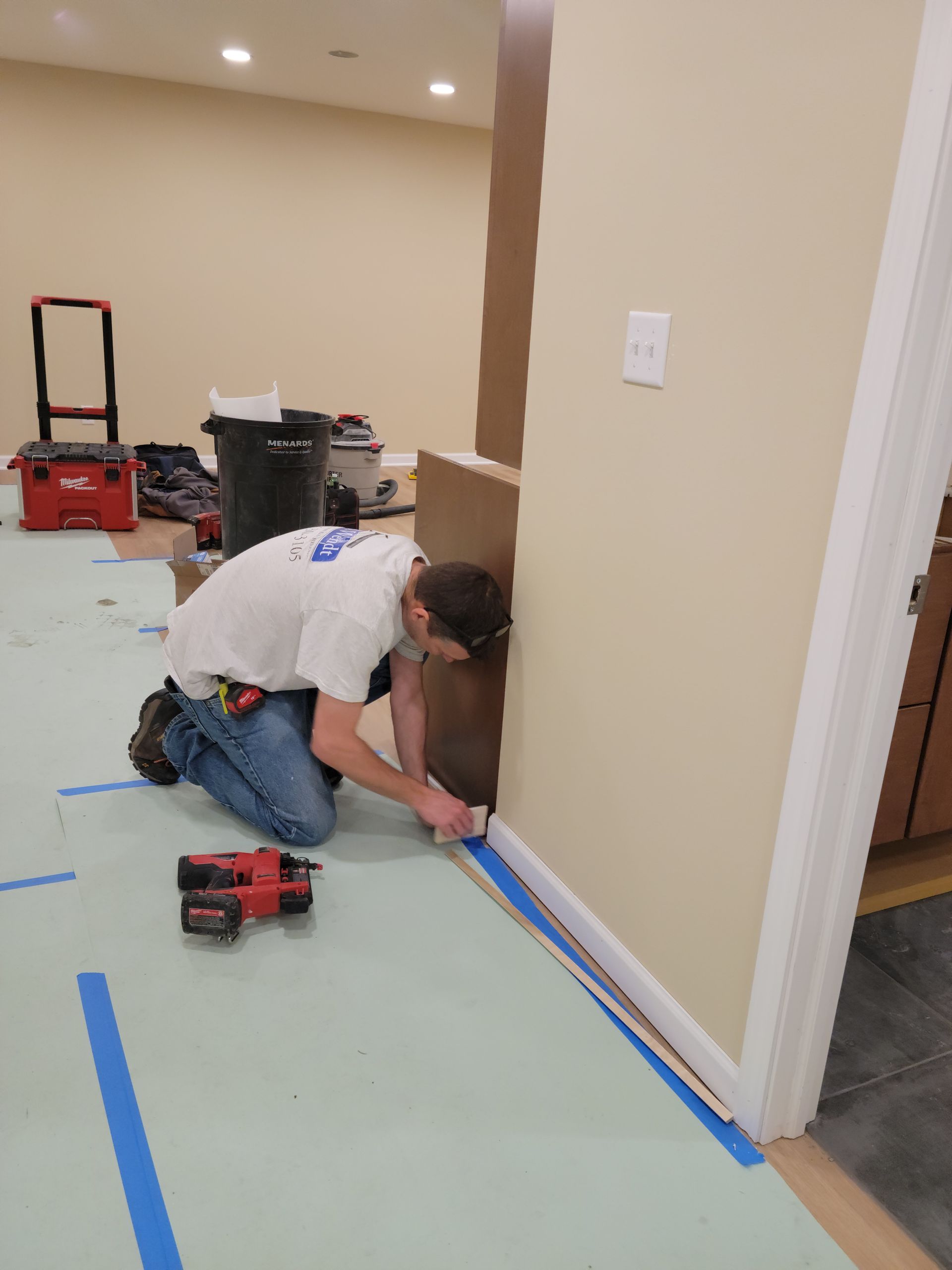 Person kneeling and measuring a floor corner with blue tape and a red power tool in a room under renovation