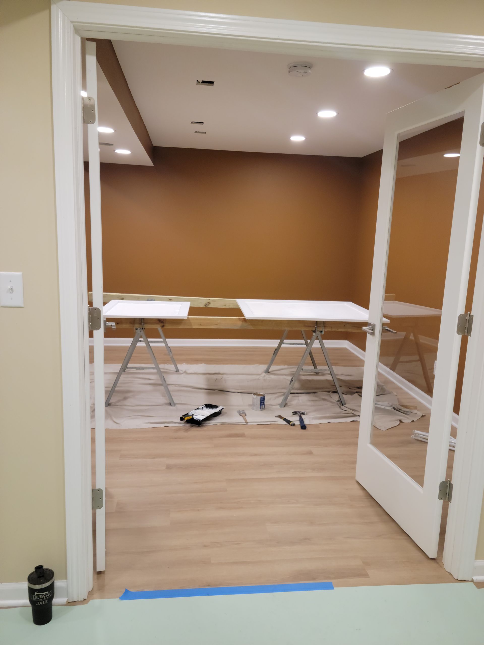 Empty room with folding tables, tan walls, and open double doors on a wood floor.