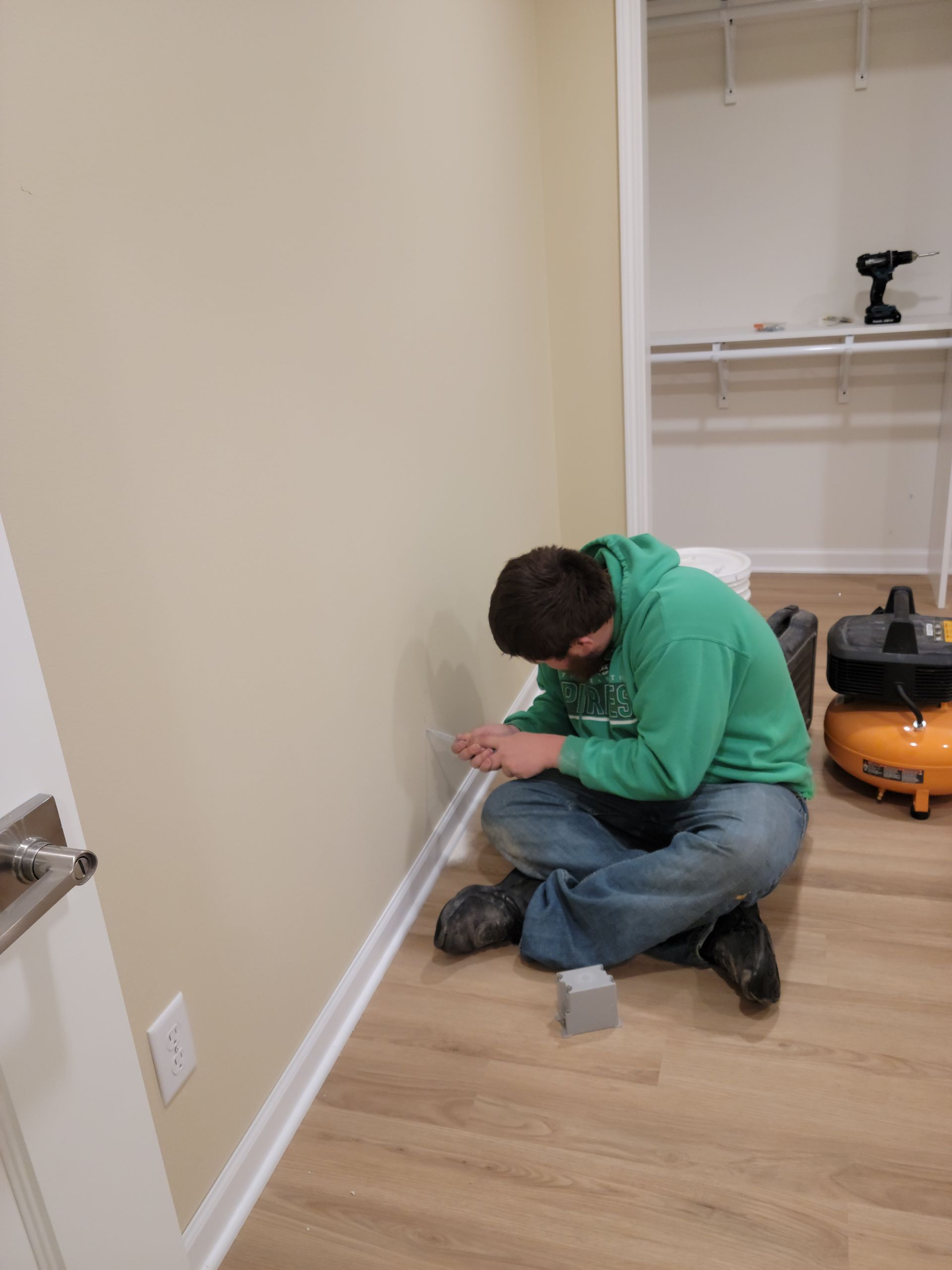 Two people kneel by a wall, working together on a baseboard in an empty room.