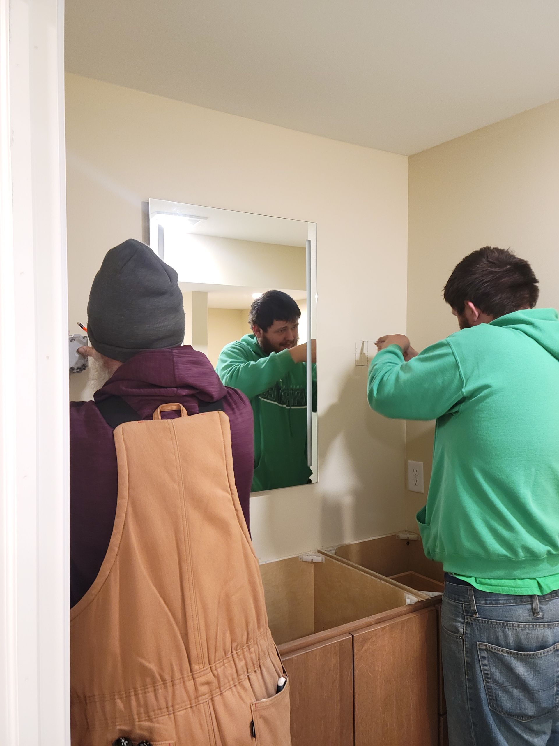 Two people in a bathroom hanging a mirror above a sink and cabinet.