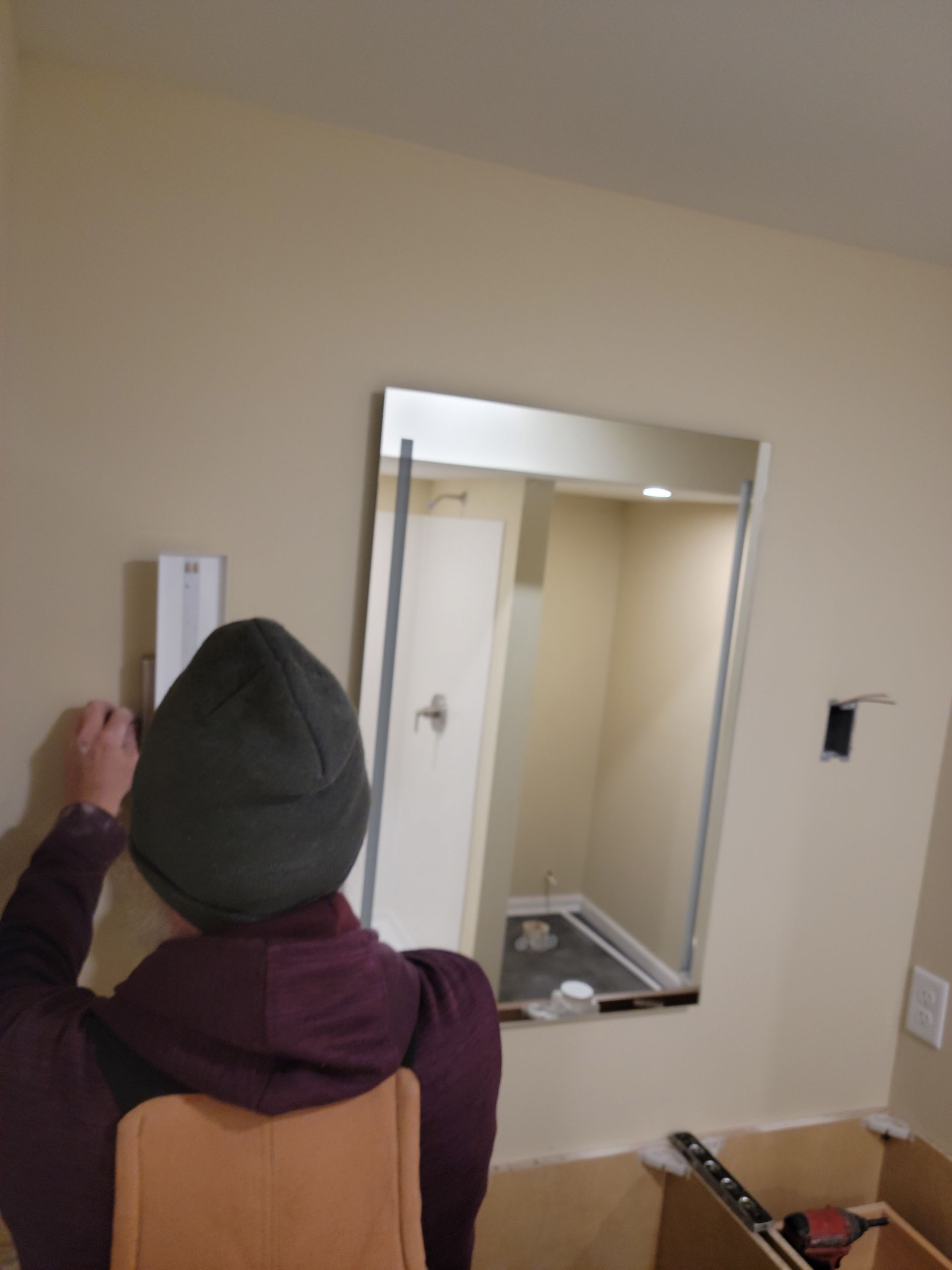 Person in a beanie installing a wall thermostat in a beige hallway near a mirror.