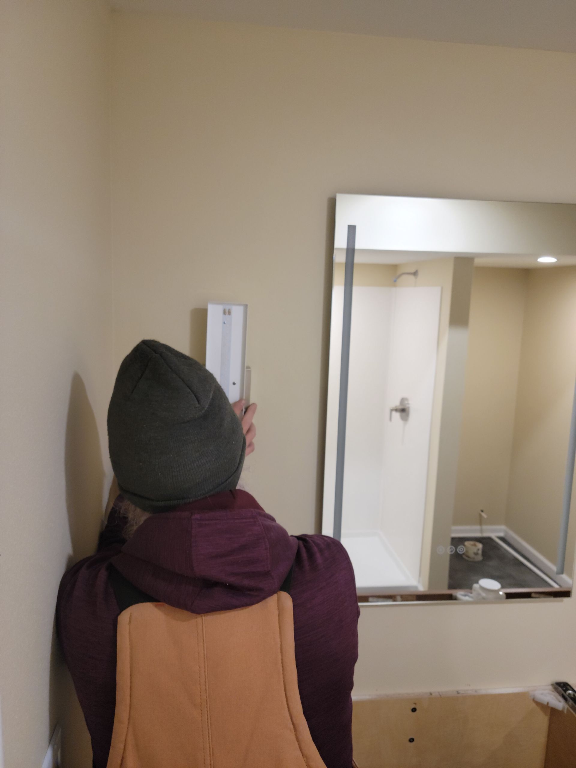 Person in winter hat and scarf looking into a wall mirror in a beige hallway.
