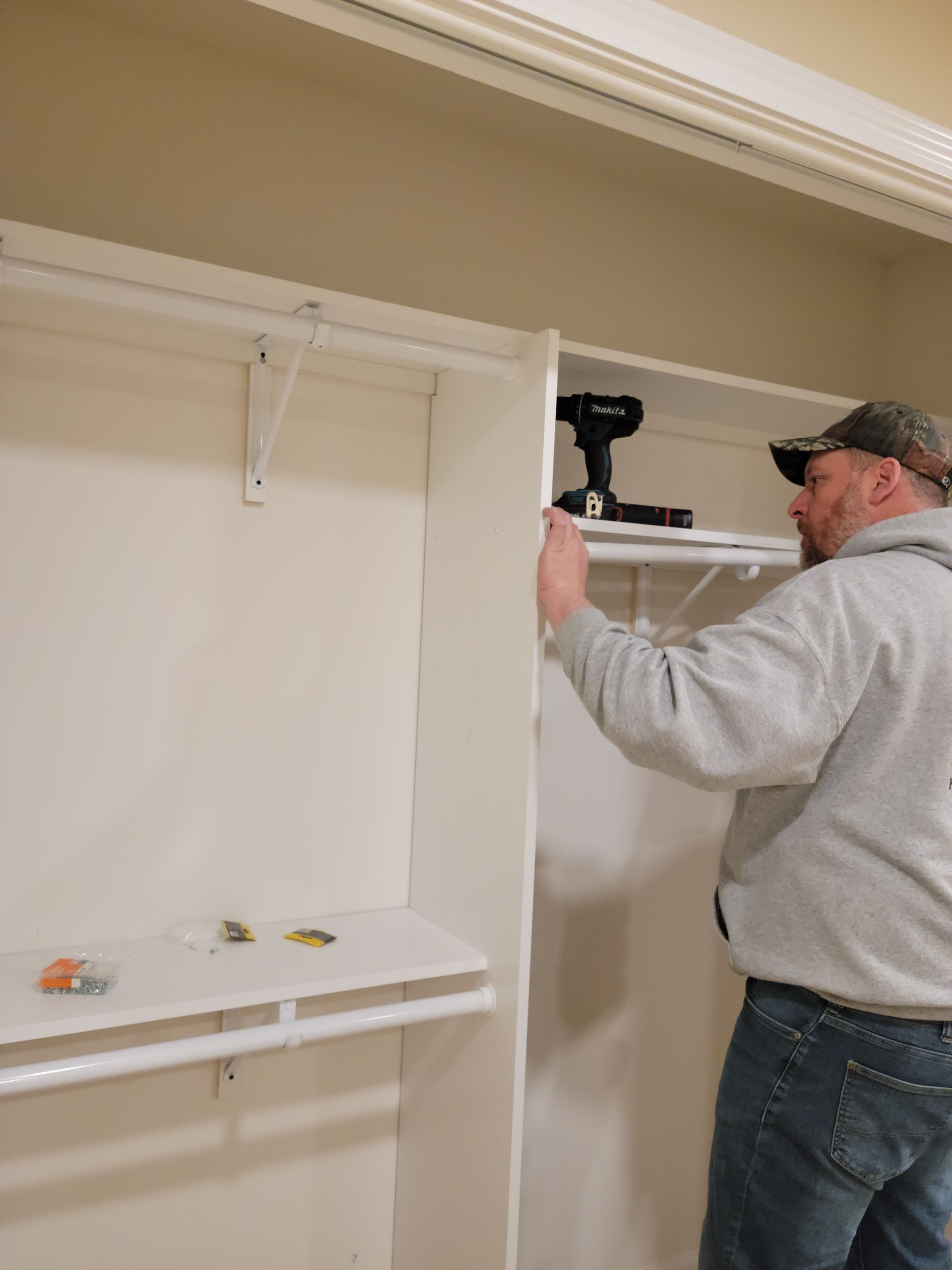 Man installing a white closet shelf with a power drill in a beige room