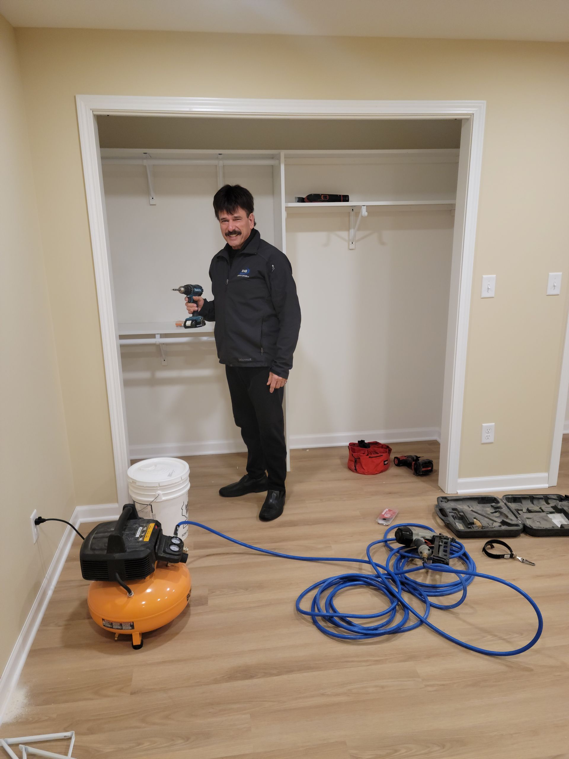 Worker standing in an empty closet beside an orange air compressor and coiled blue hose