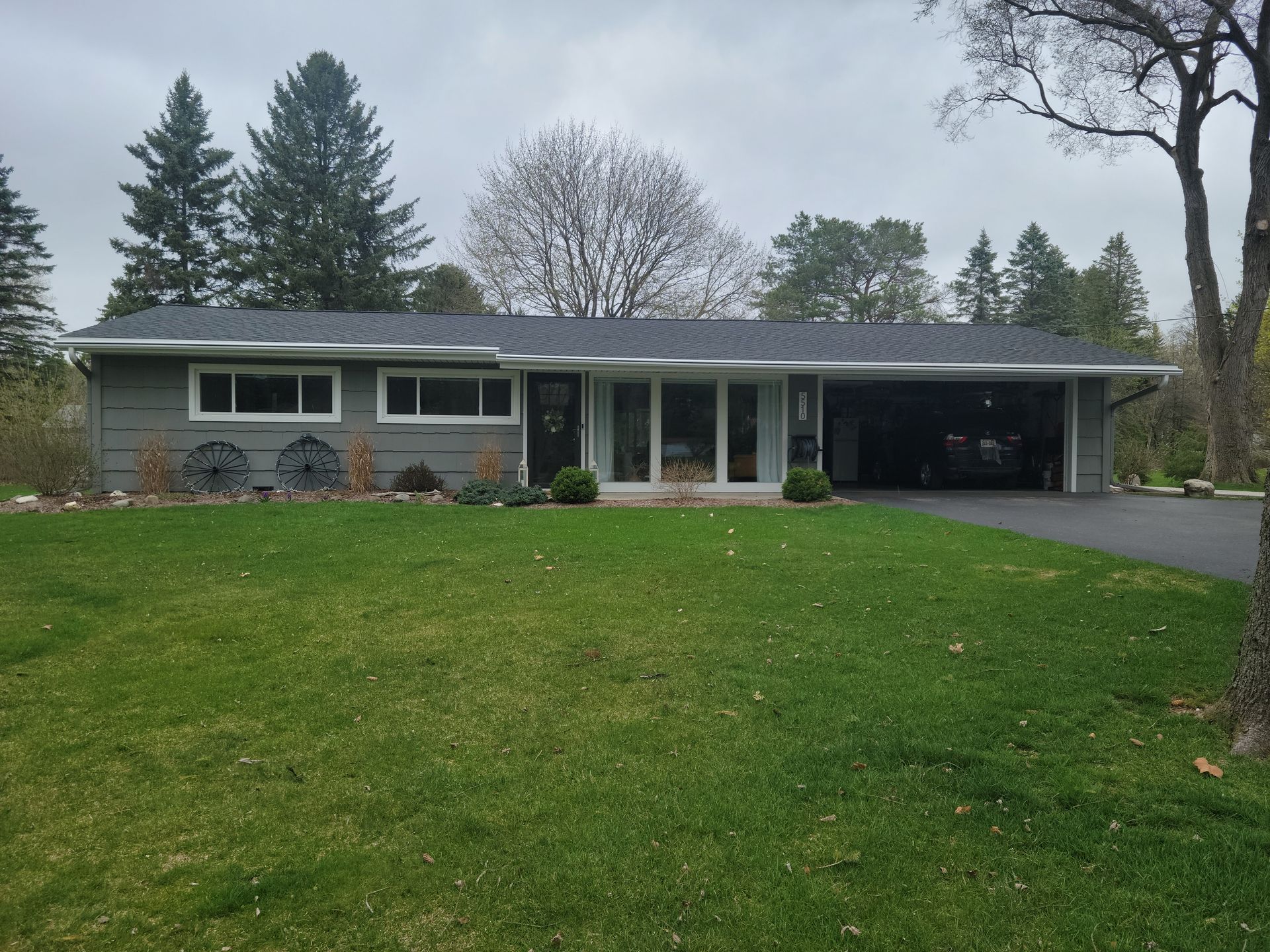 Single-story gray house with a large green lawn, driveway, and trees under an overcast sky