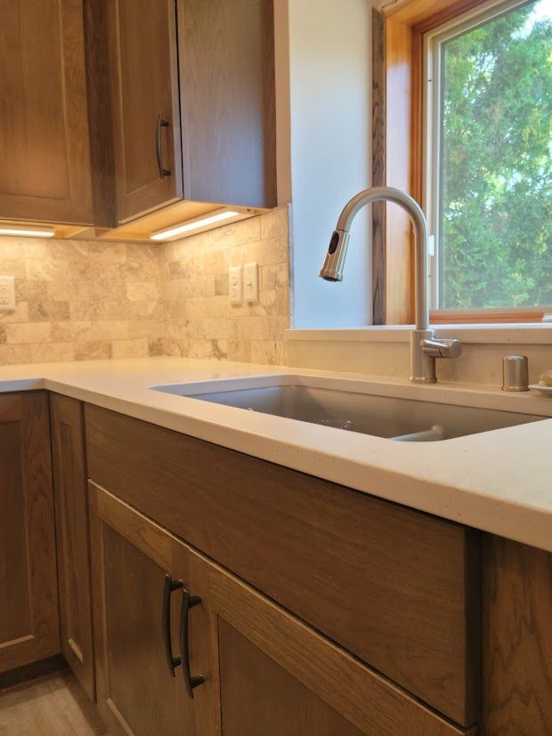 Kitchen sink with chrome faucet, beige countertop, wood cabinets, and a window with greenery outside