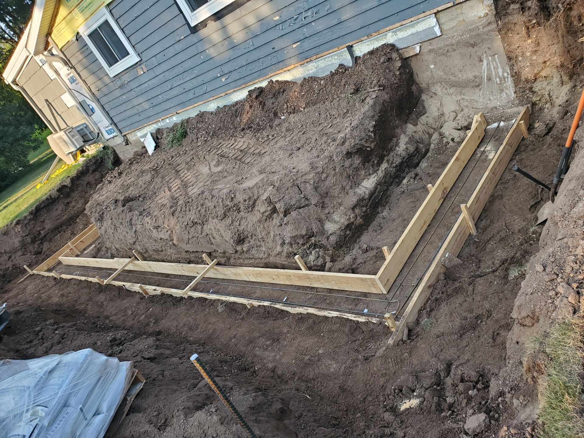 Foundation work beside a house, with concrete forms and excavated soil in a muddy yard.