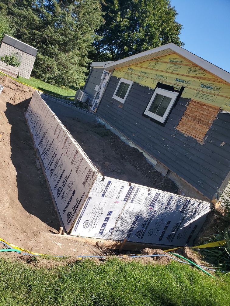 Tilted house with exposed foundation and wrapped exterior under construction on a sloped lot.