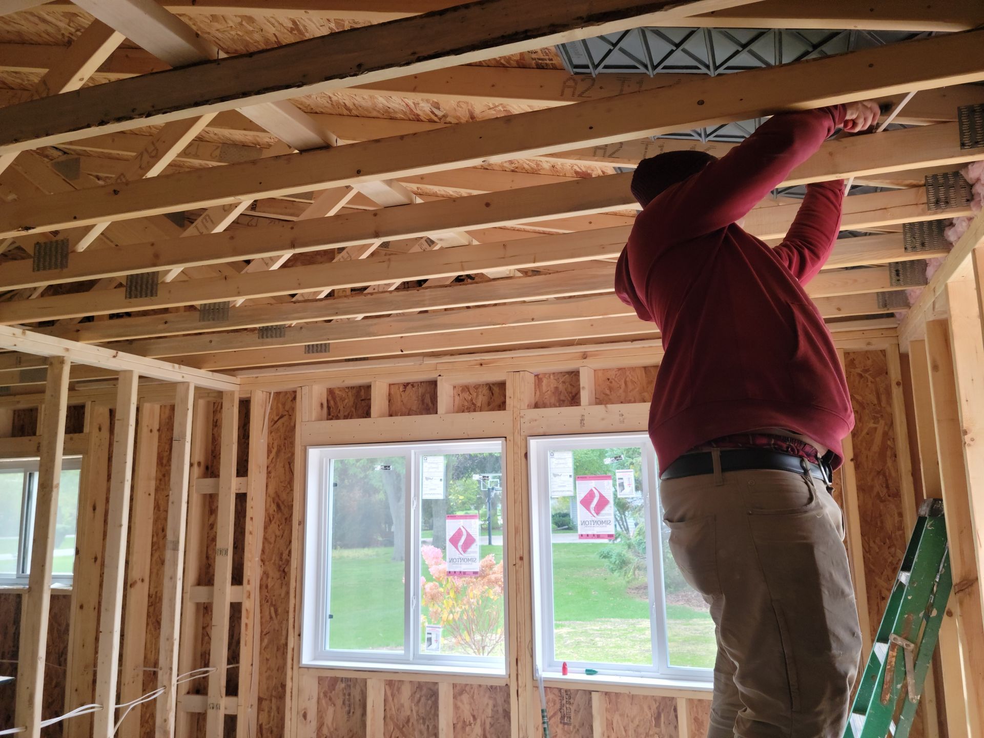 Person working on exposed ceiling framing in a room under construction, with a window and ladder nearby