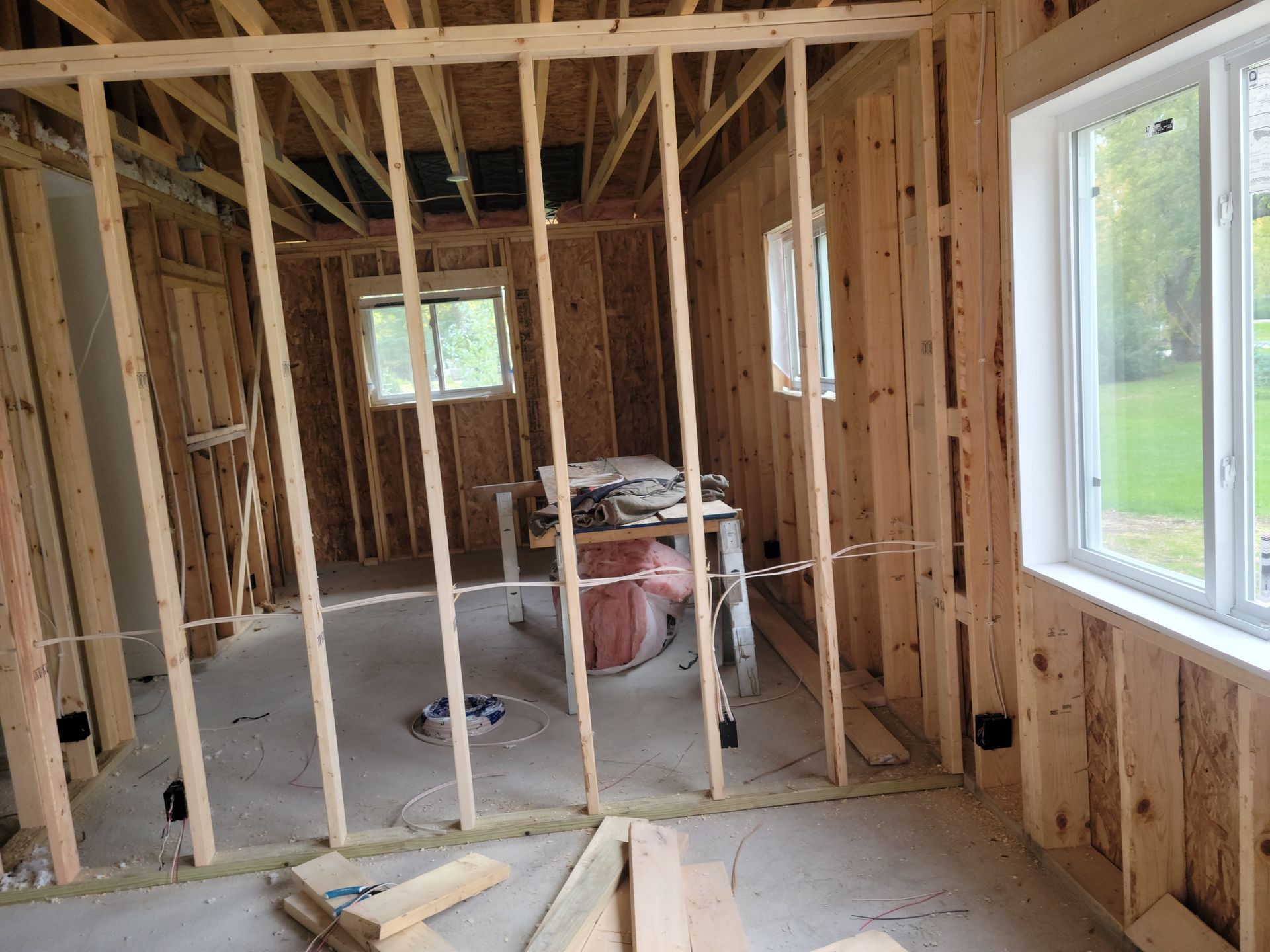 Unfinished interior room with exposed wooden framing, insulation, and a large window showing green lawn outside.
