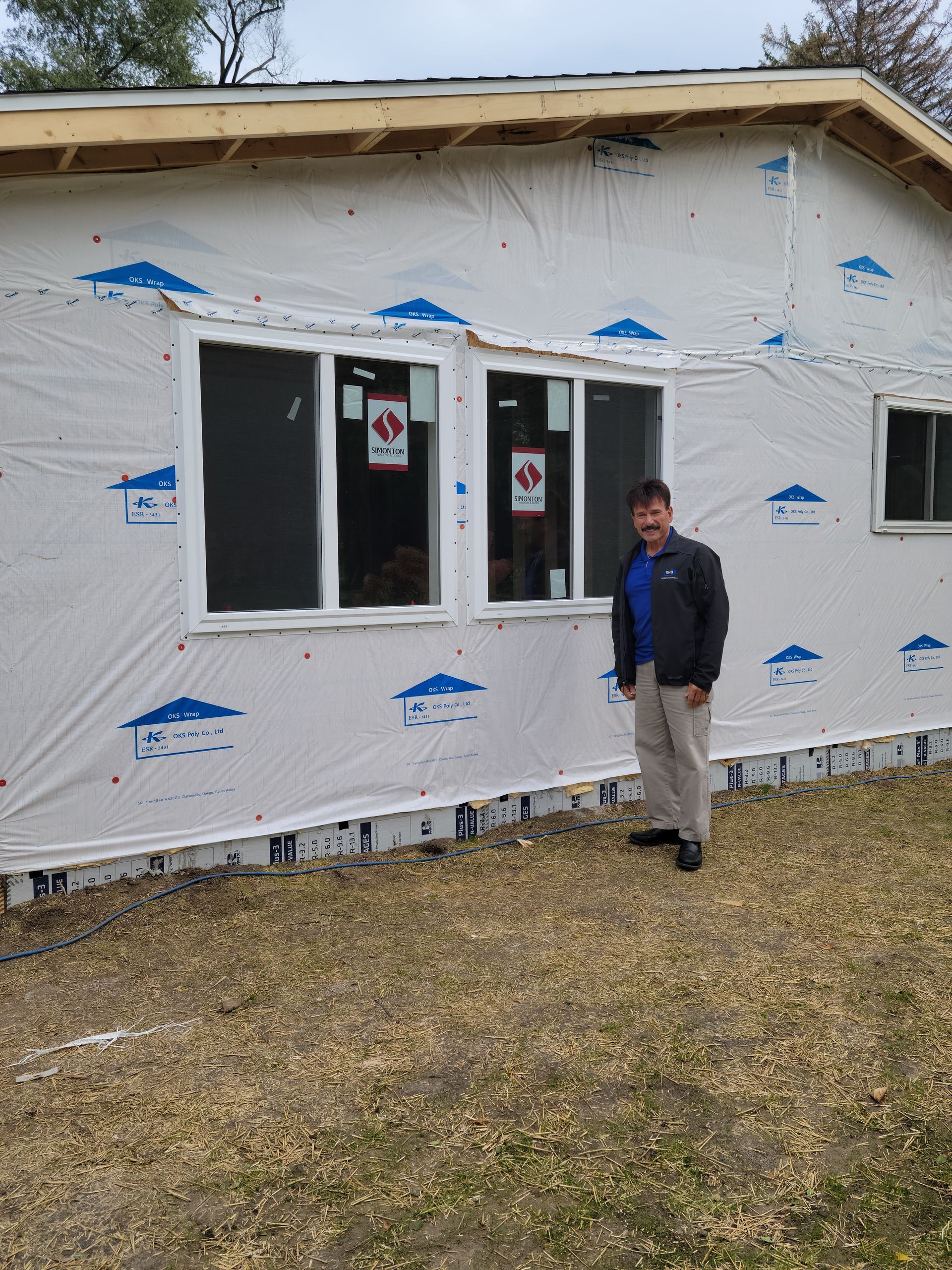 Person standing outside a partially finished house wrapped in white house wrap with blue tape.