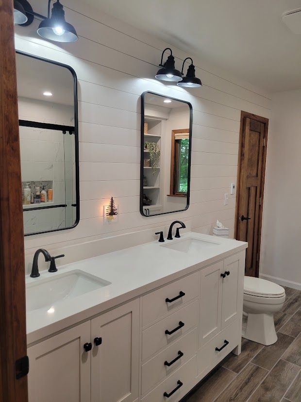 Bright bathroom with double vanity, black fixtures, white shiplap walls, and a toilet by the wooden door.
