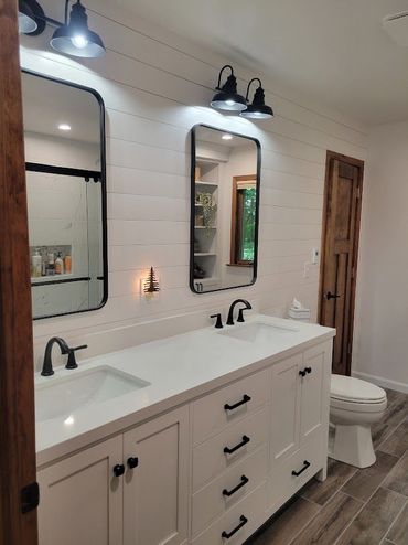 Bright bathroom with double vanity, black fixtures, white shiplap walls, and a toilet by the wooden door.