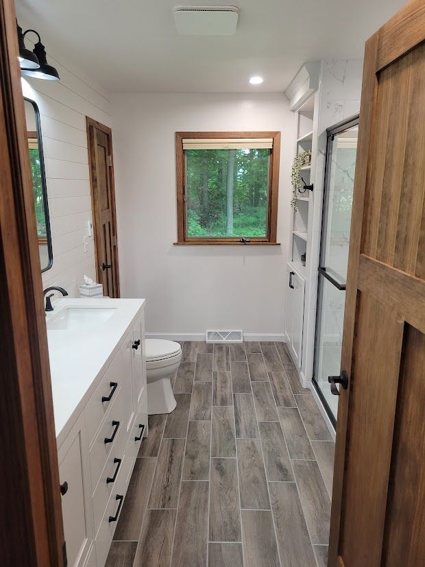 Narrow bathroom with white vanity, toilet, wood door, gray tile floor, and window at the far end