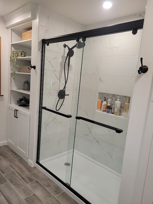 Glass shower enclosure with black hardware in a modern bathroom, next to open shelving and gray tile floor
