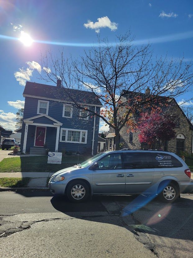 Sunny street with silver minivan parked in front of a two-story house and leafless tree.