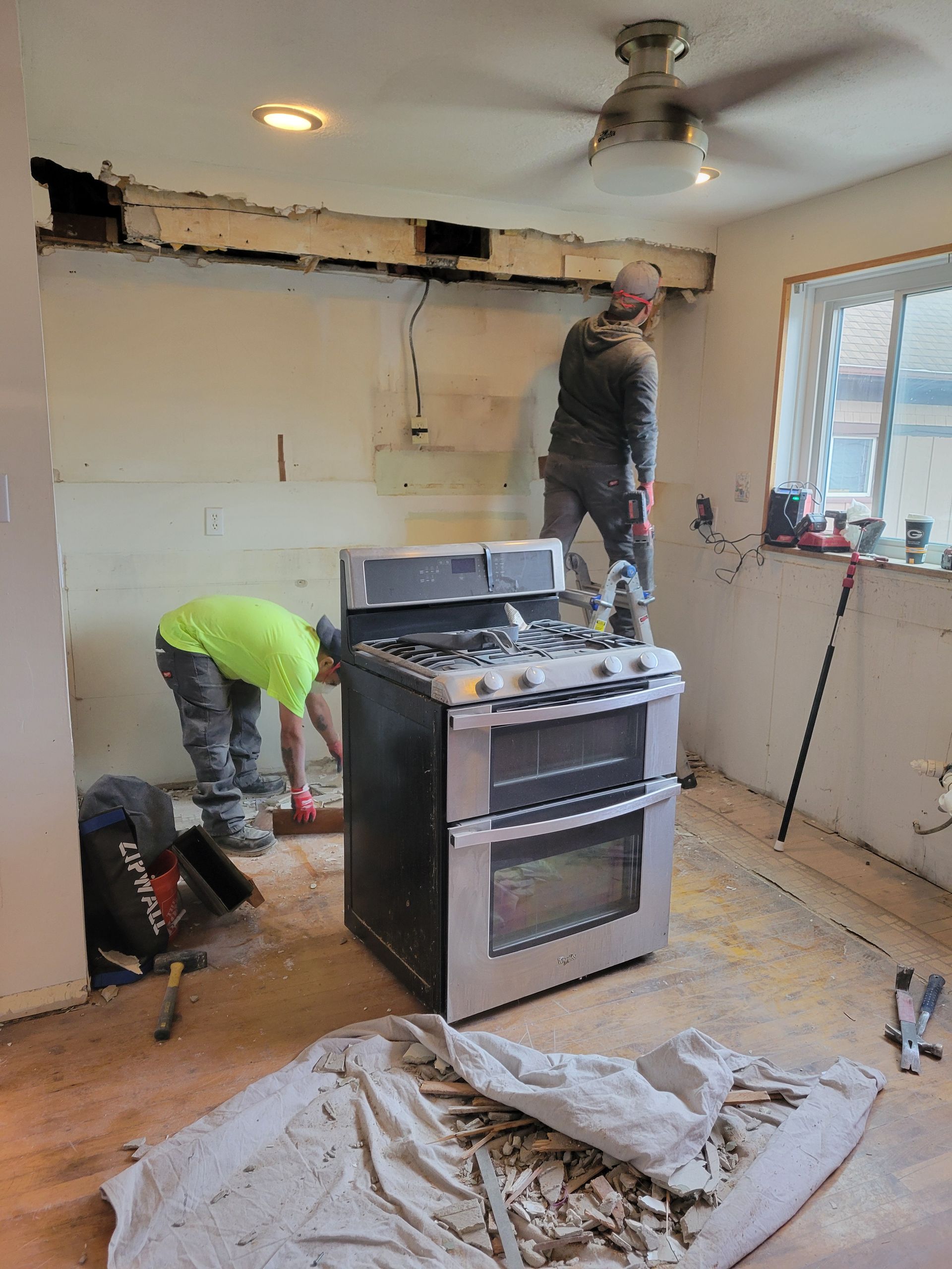Two workers renovating a kitchen, with a stove in the center and a damaged ceiling under repair